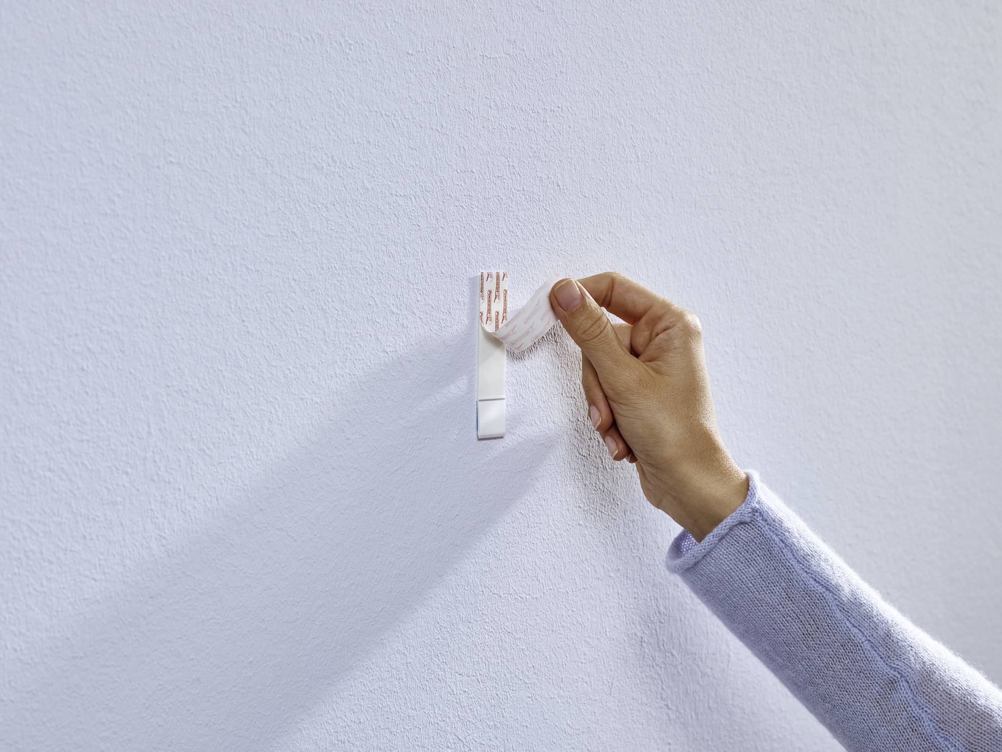 Une main fixe un crochet adhésif sur un mur blanc. Le crochet est doucement pressé pour l'installer fermement.