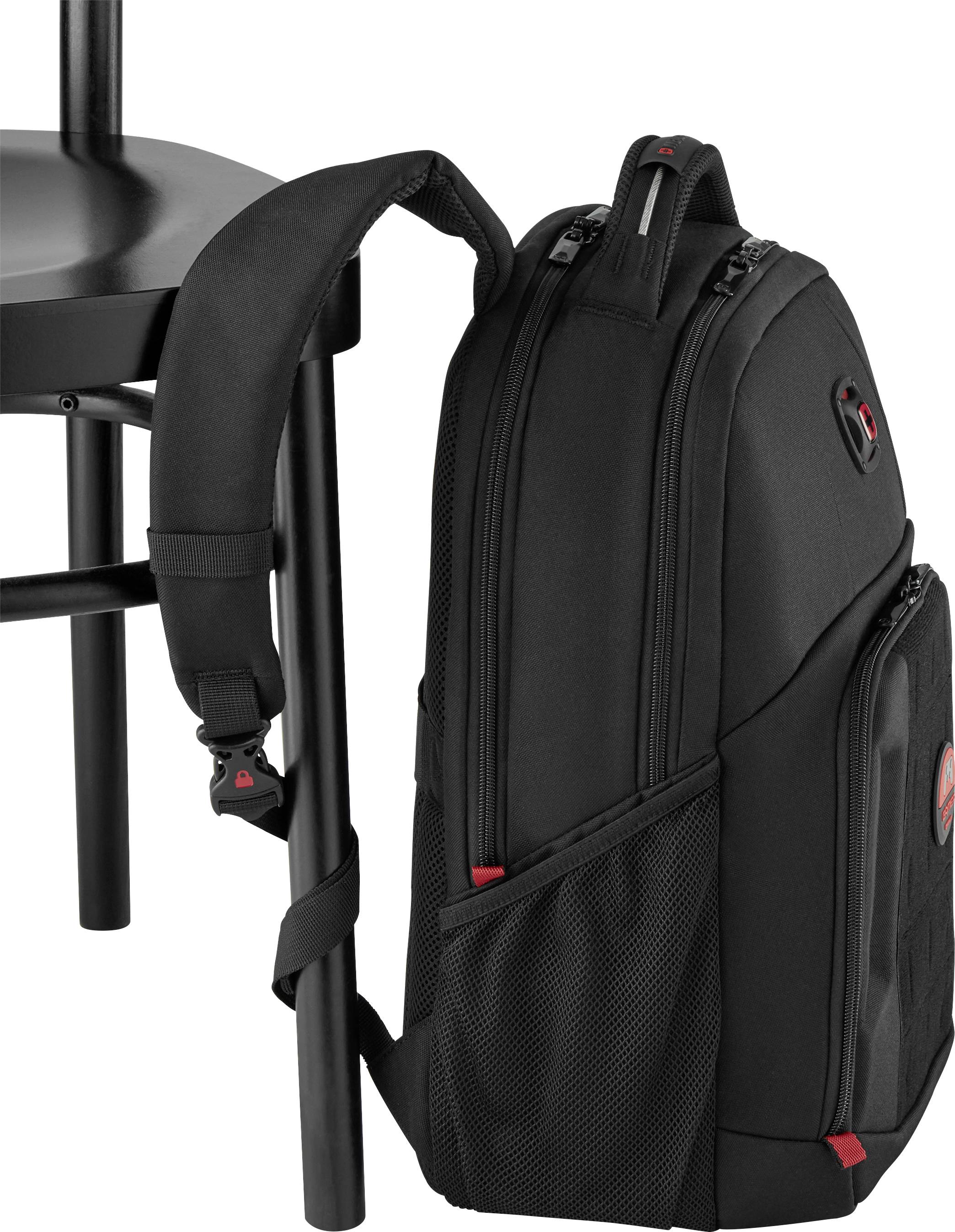 Un sac à dos noir est appuyé contre une chaise noire. Il présente des accents rouges et plusieurs fermetures à glissière.