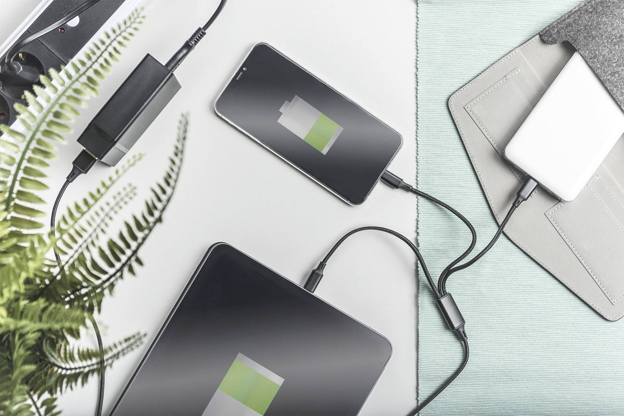 Une tablette et un smartphone sont en train de se charger simultanément ; tous deux affichent un symbole de batterie partiellement chargé. Un chargeur et un bloc-notes sont visibles à côté des appareils.