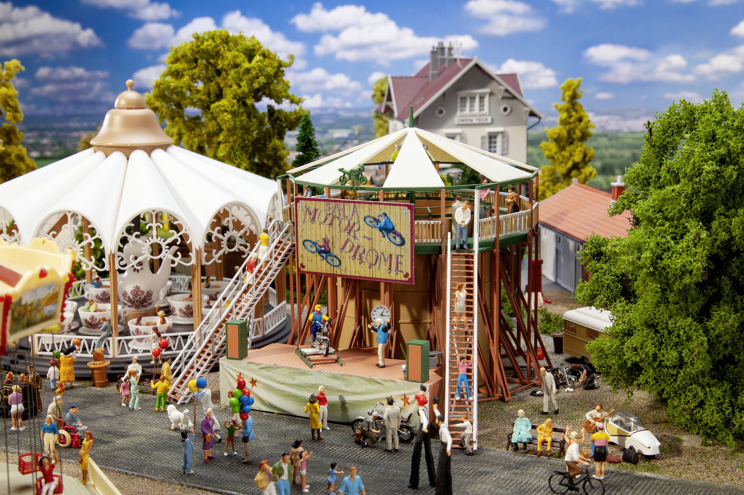 Un parc de loisirs miniature avec un carrousel, une scène et des personnes. Sur la scène, une affiche portant l'inscription « My Place or My Chrome ».