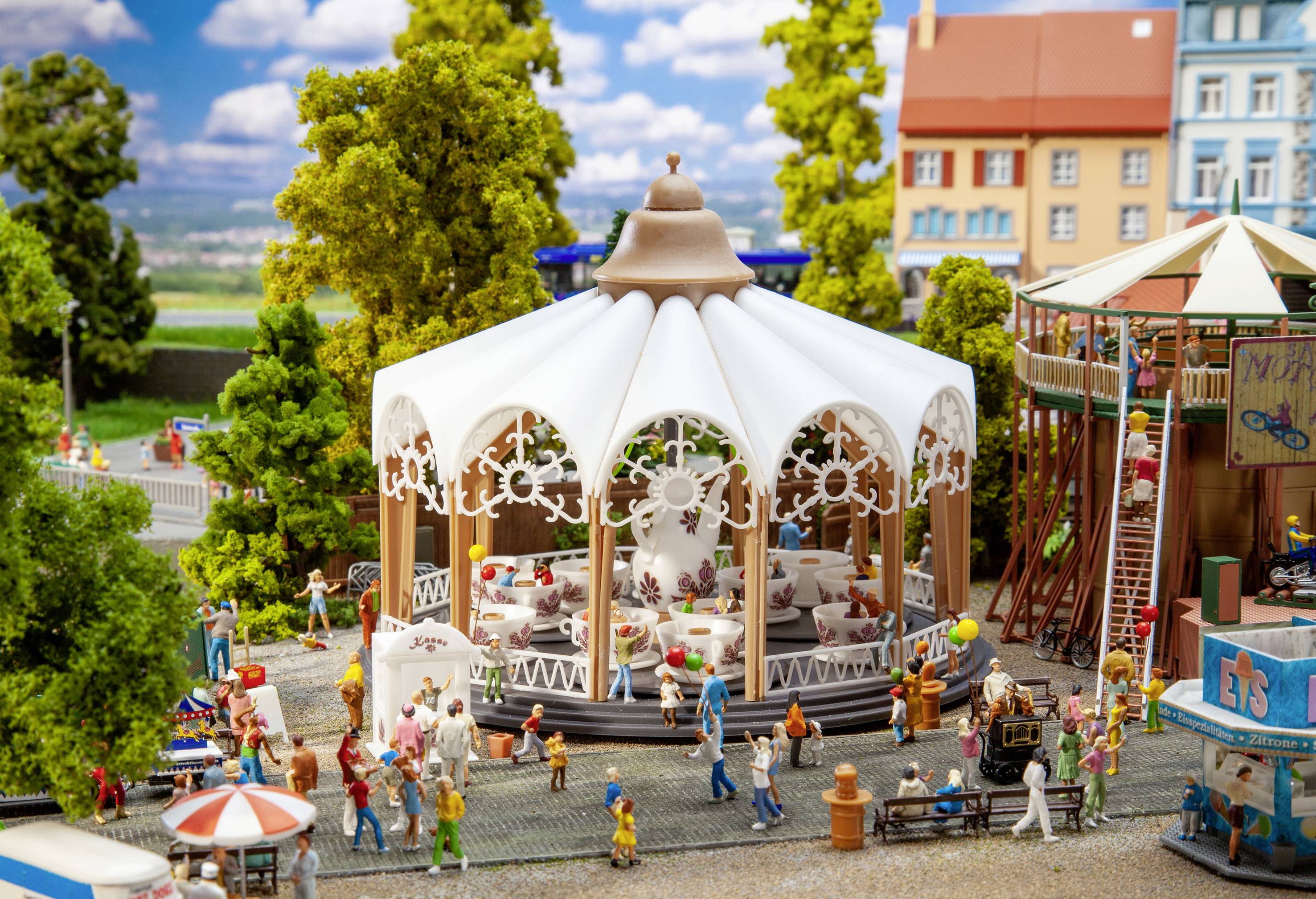 Petit monde miniature avec un pavillon de carrousel, entouré de personnages et d'arbres, dans le parc municipal par une journée ensoleillée. Des enfants et des adultes interagissent.