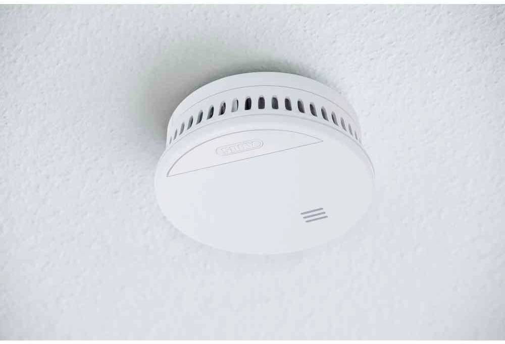 Un détecteur de fumée blanc est fixé au plafond. Il possède des fentes d'aération sur le côté et un design simple et rond.
