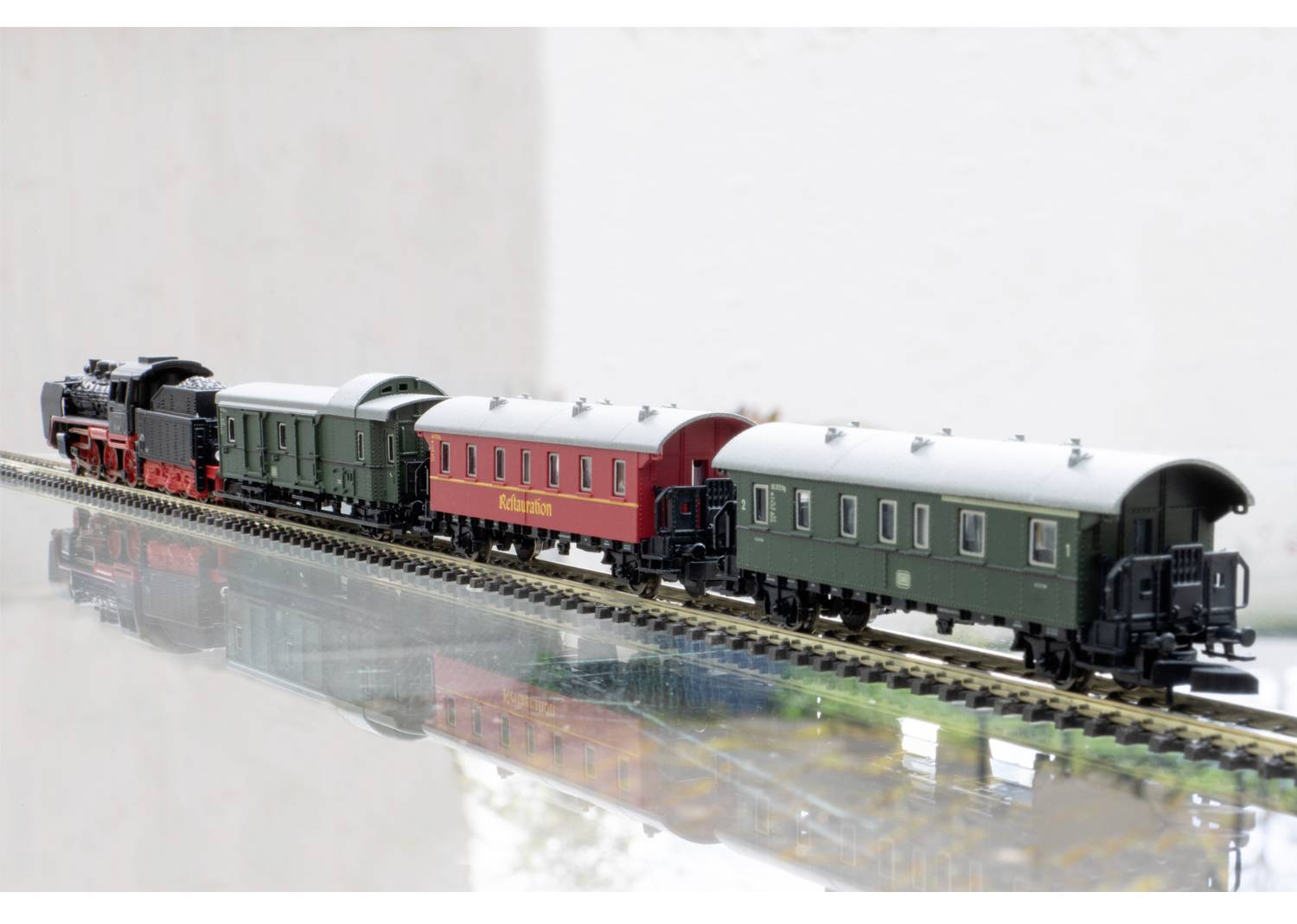 Un train miniature avec une locomotive à vapeur et quatre wagons sur une voie ferrée. Deux wagons sont verts, un est rouge avec l'inscription 'Märklin'.