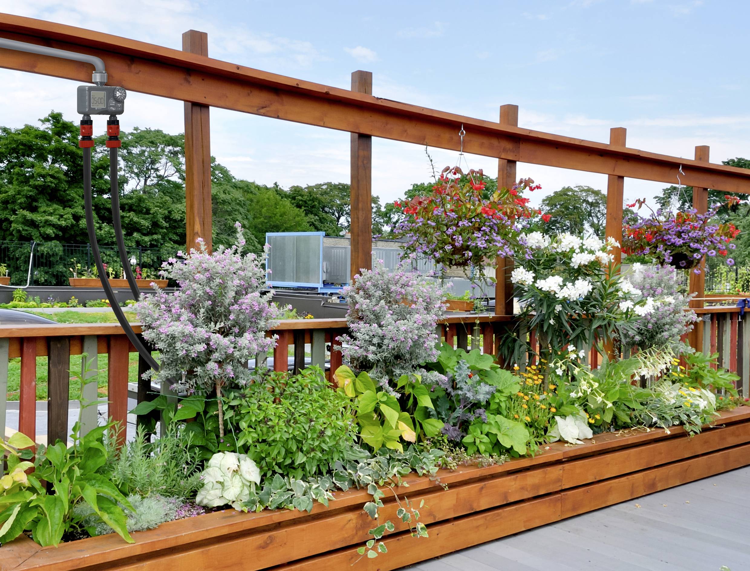 Un carré de jardin surélevé rempli de fleurs colorées et de feuillage vert est installé sur une terrasse. En arrière-plan, on aperçoit des arbres et un ciel bleu.