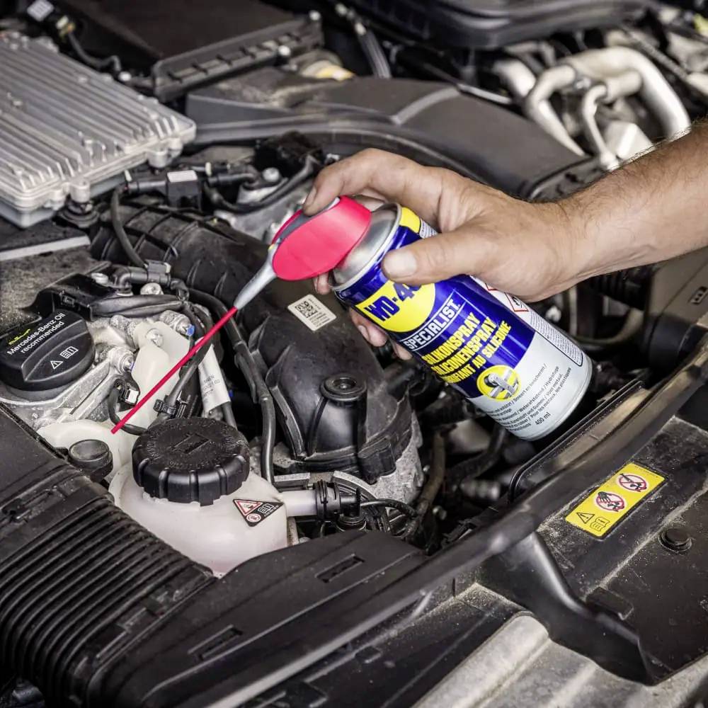 Une main pulvérise un lubrifiant sur un moteur de pièce automobile afin d'effectuer une maintenance. Le compartiment moteur est clairement visible.