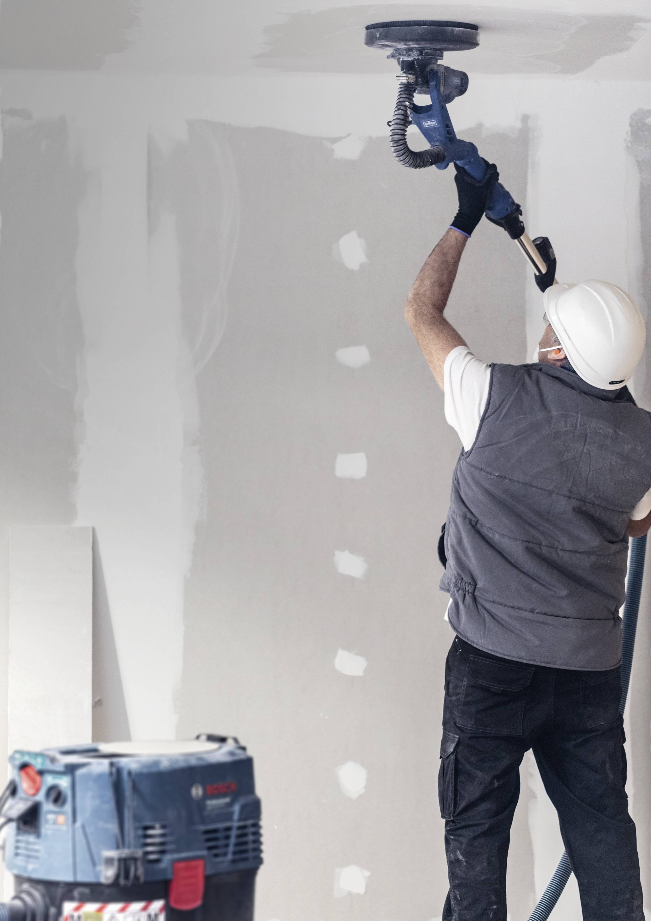 Un homme ponce un mur dans une pièce, portant des vêtements de protection et utilisant une ponceuse. Signes de rénovation ou de travaux en cours.