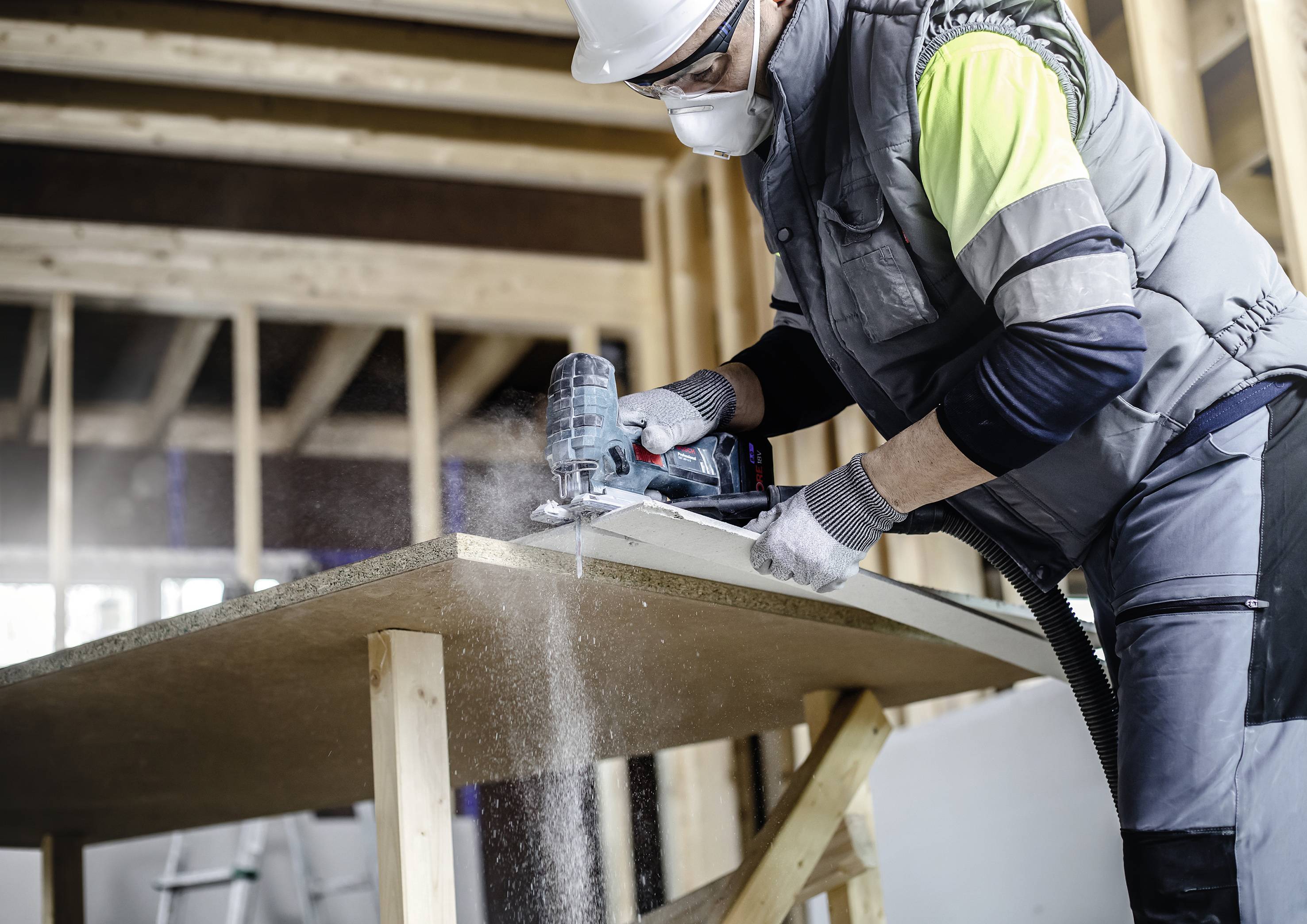 Un ouvrier porte des vêtements de protection et coupe du bois avec une scie électrique dans un bâtiment en construction.