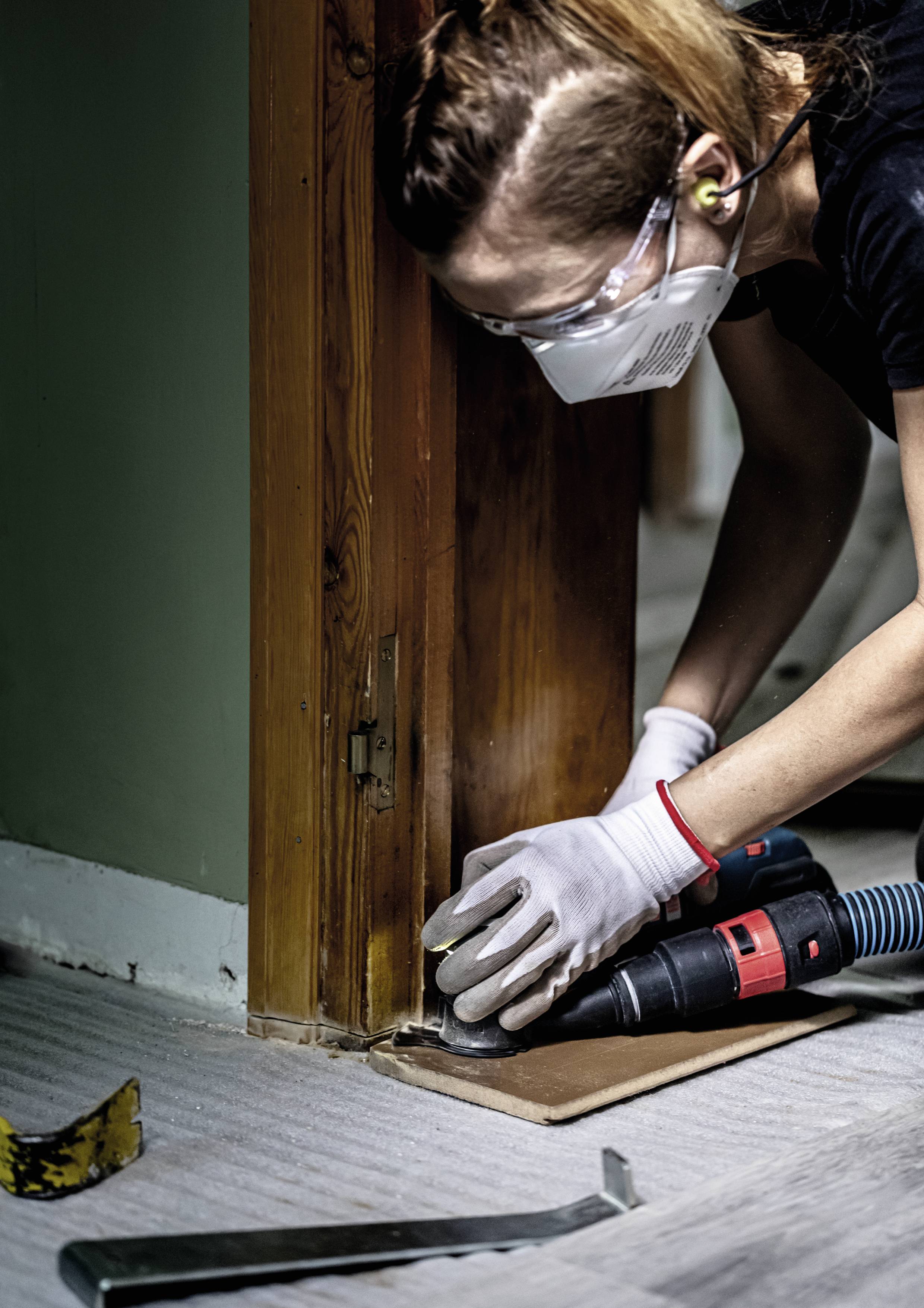 Une personne porte des lunettes de protection et un masque et utilise un outil électrique pour travailler le bois sur un chambranle de porte.