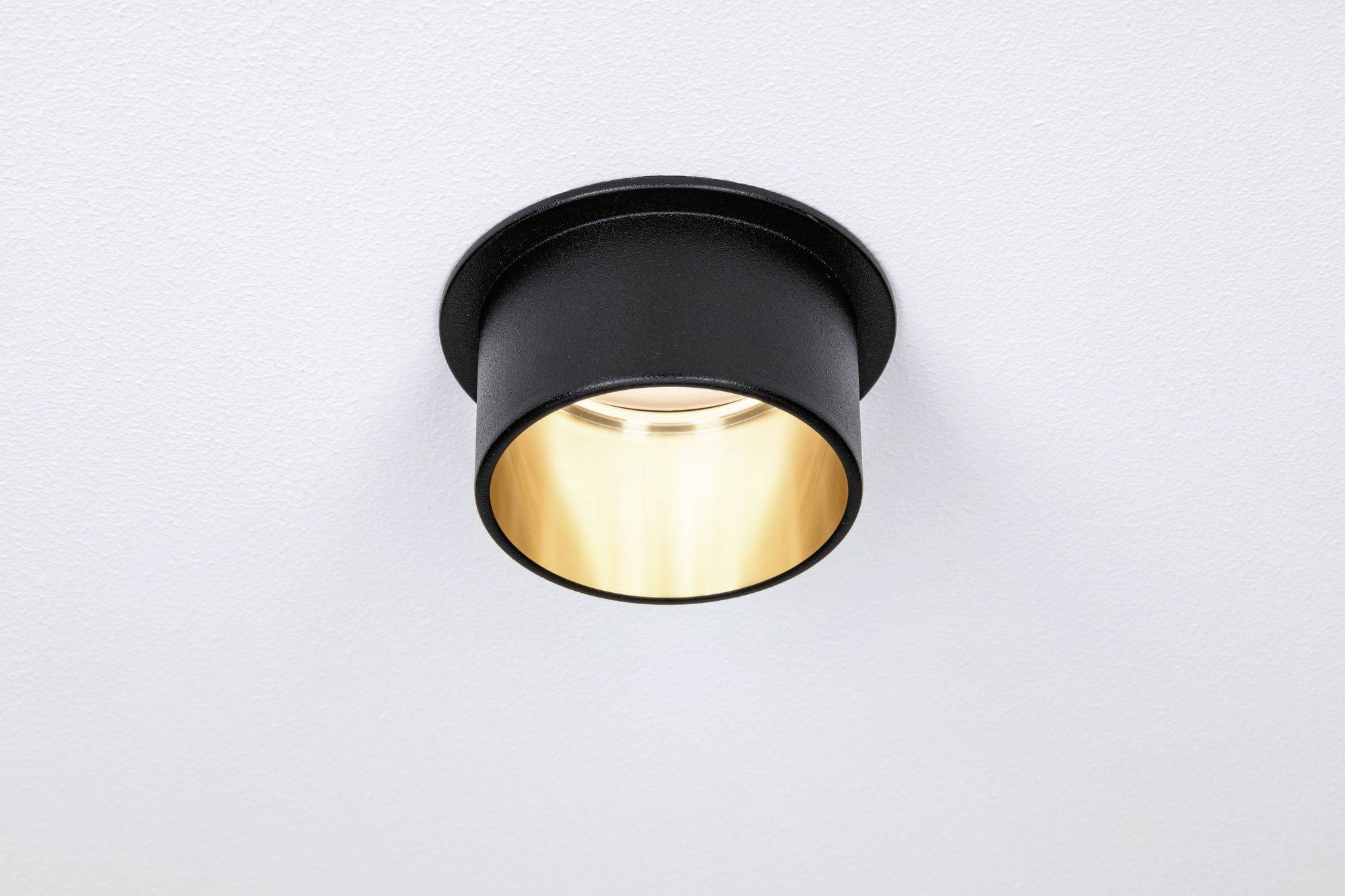 Un plafonnier rond noir avec une lumière chaude, monté sur un plafond blanc. Il éclaire directement vers le bas.