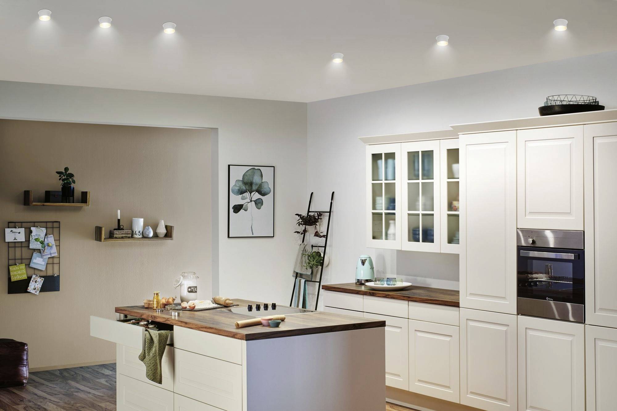 Cuisine lumineuse et moderne avec des armoires couleur crème, un îlot en bois et un four. Des étagères et de la décoration sont accrochées au mur.