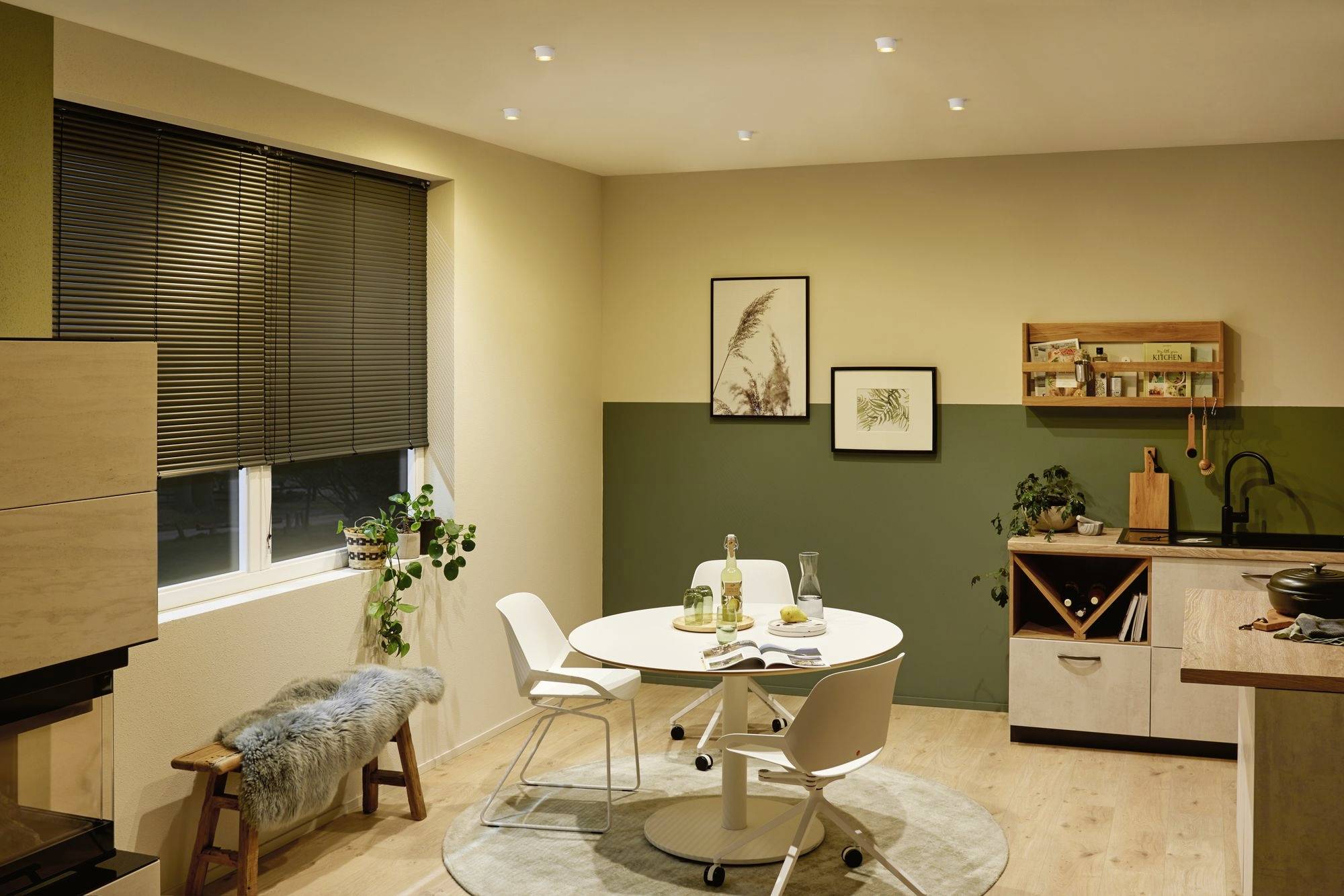 Une salle à manger élégamment aménagée avec une table ronde et quatre chaises. Des tableaux sont accrochés au mur, des plantes sont disposées sur le rebord de la fenêtre et sur le sol.