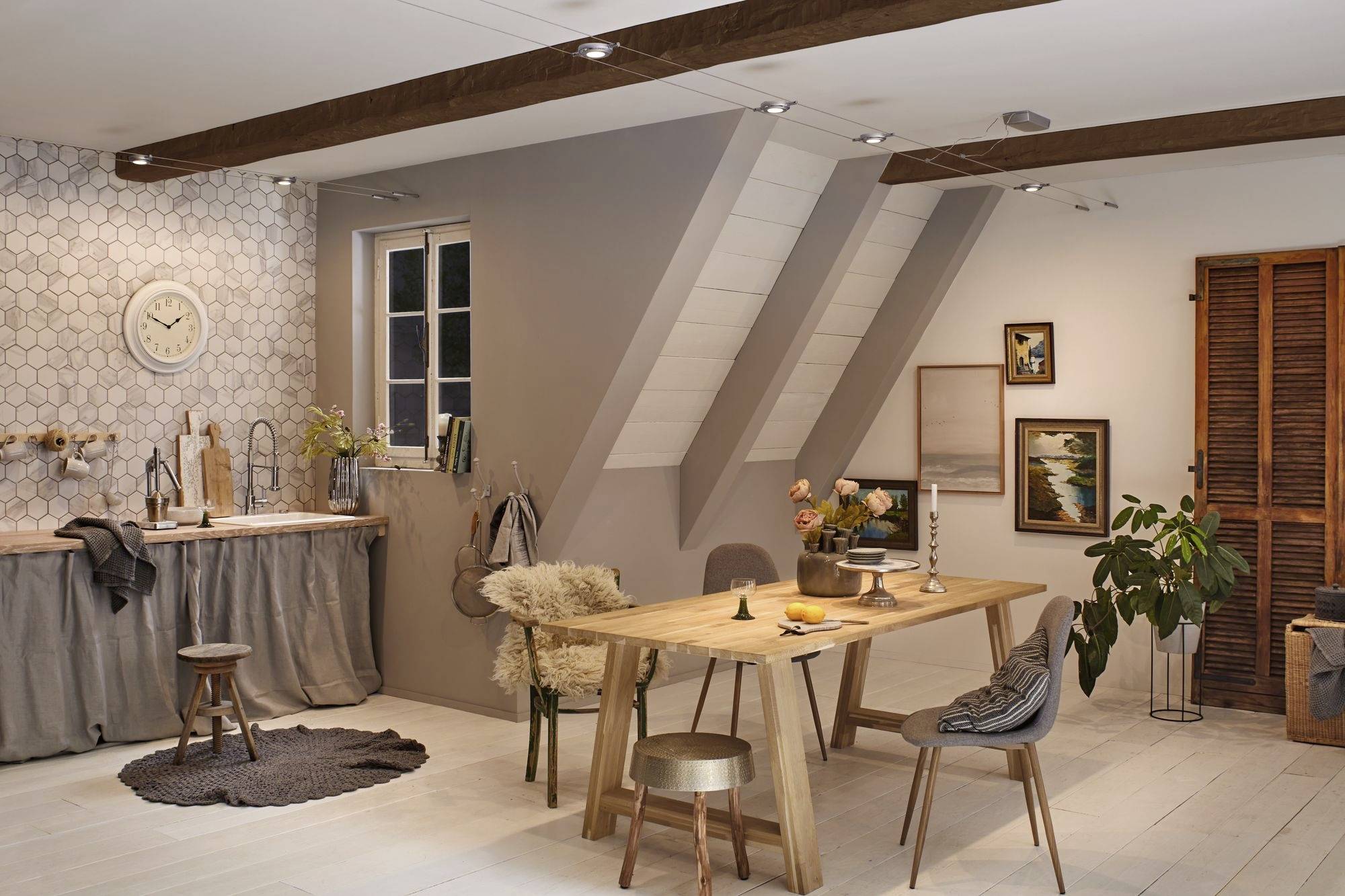 Cuisine-salon chaleureuse sous les combles avec des poutres en bois, une table en bois, des chaises, des plantes et une fenêtre. Mur carrelé avec une horloge.