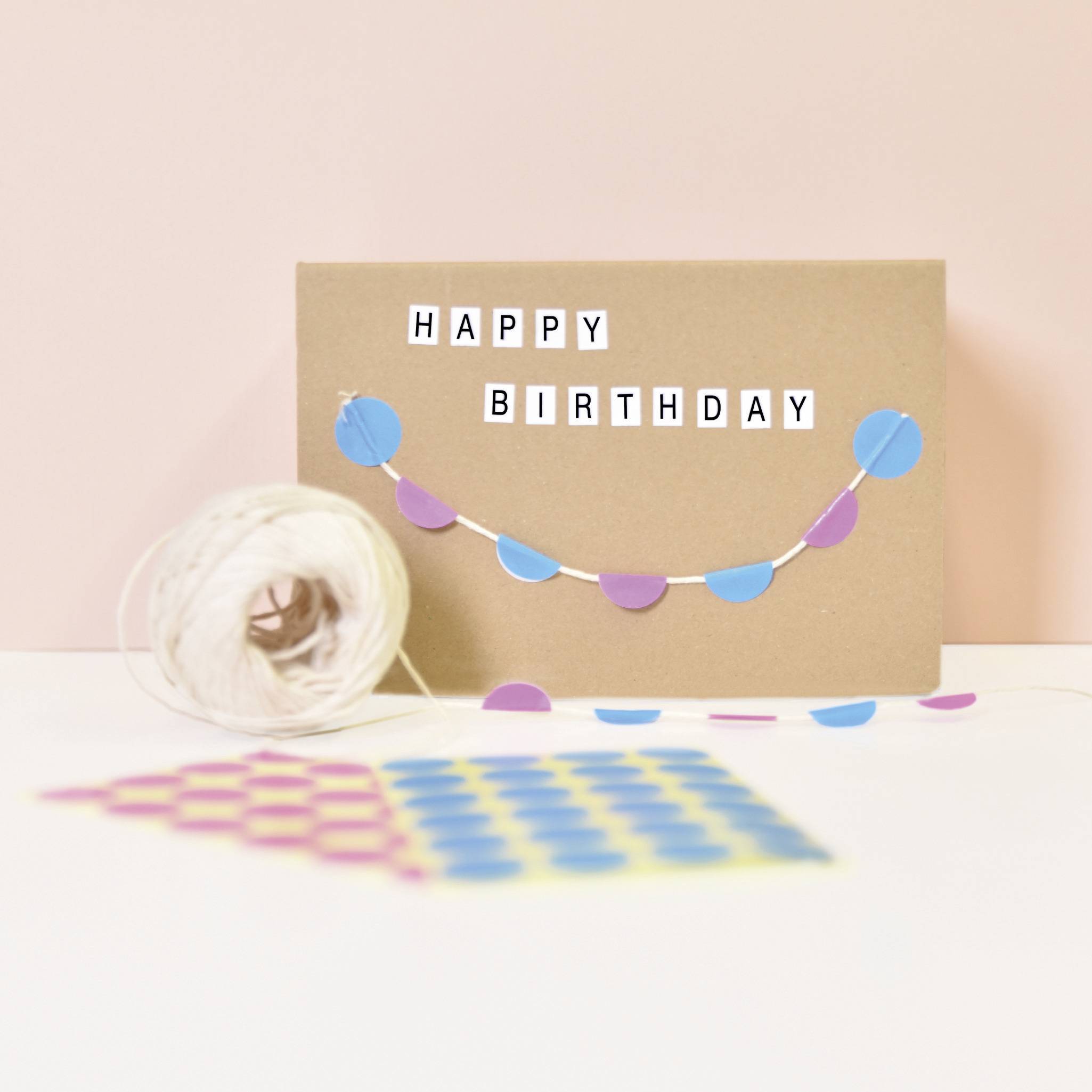 Carte d'anniversaire avec banderole colorée et inscription 'Joyeux Anniversaire' sur un fond brun, à côté de laquelle se trouve un rouleau de fil.