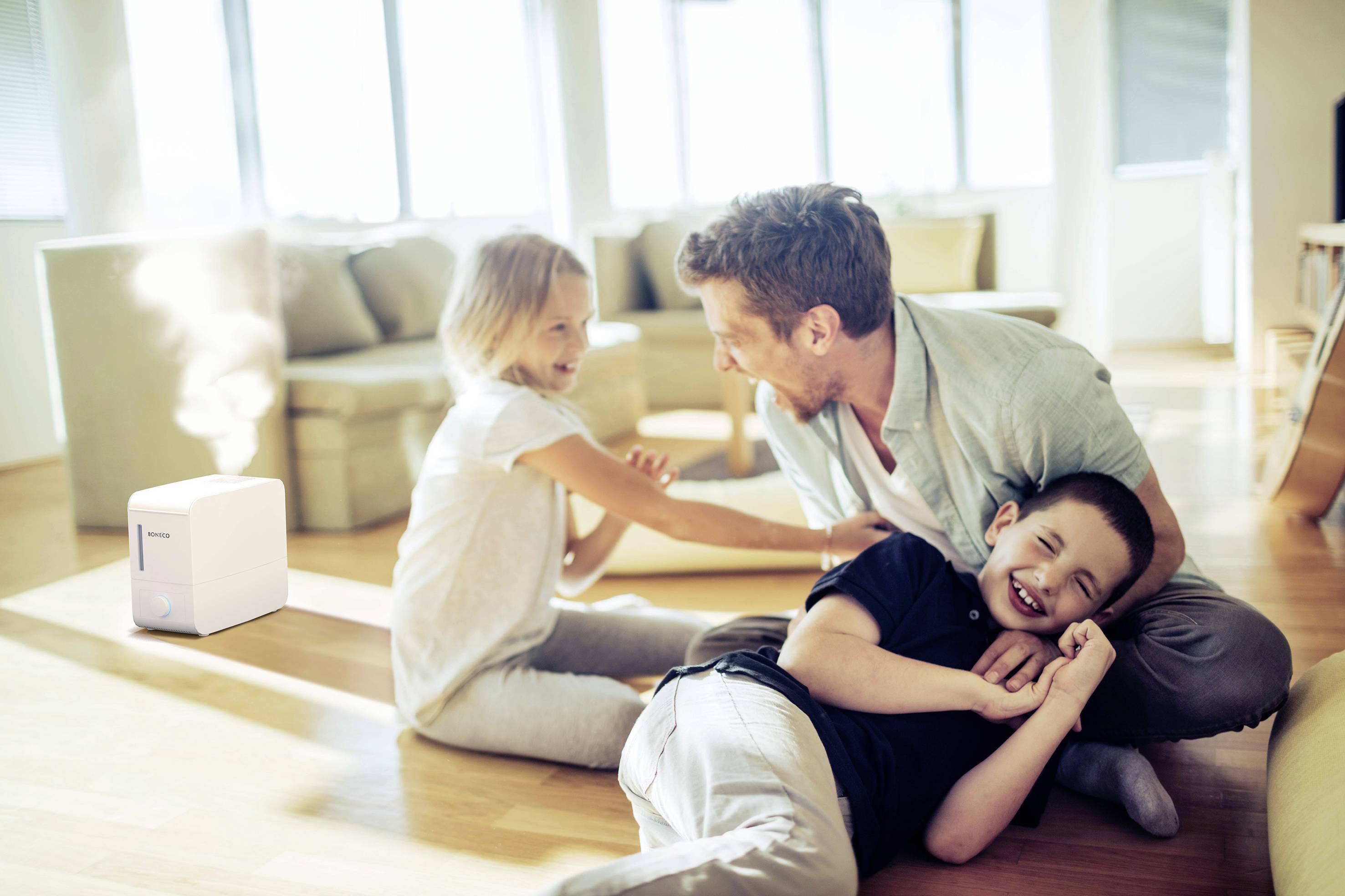 Un homme et deux enfants rient et jouent sur le sol du salon, tandis qu'un humidificateur émet de la vapeur en arrière-plan.