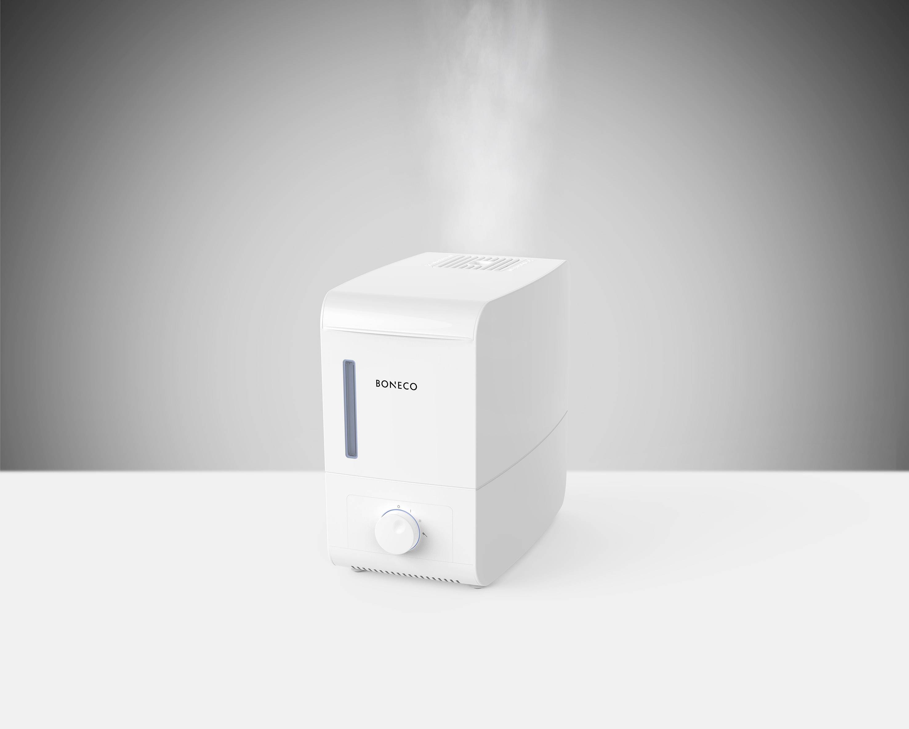 Un humidificateur blanc de Boneco, qui émet de la vapeur, est posé sur une surface blanche devant un fond gris.