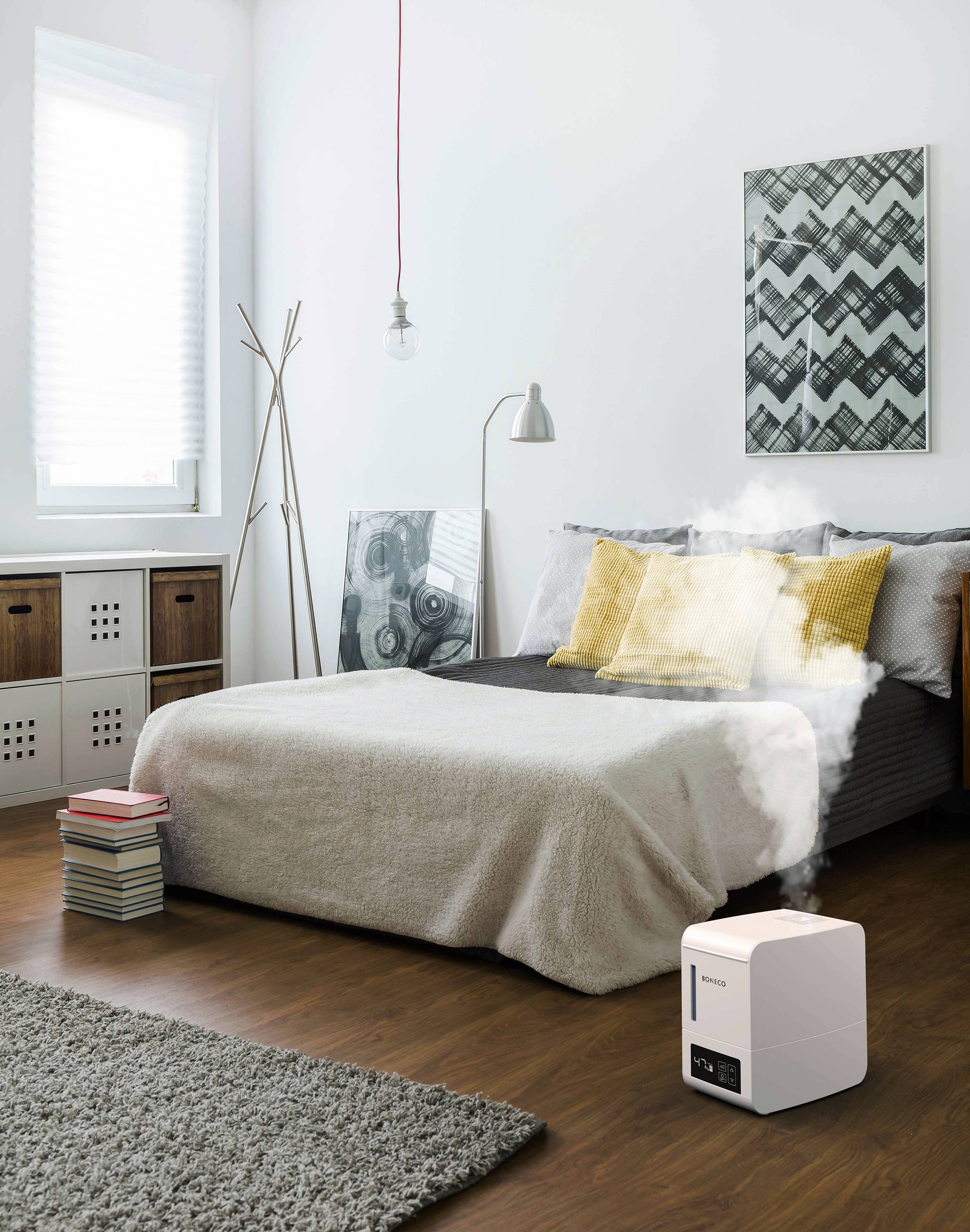 Une chambre avec un sol en bois, un lit double recouvert d'une couverture grise et de coussins jaunes. Un humidificateur d'air est placé sur le sol.