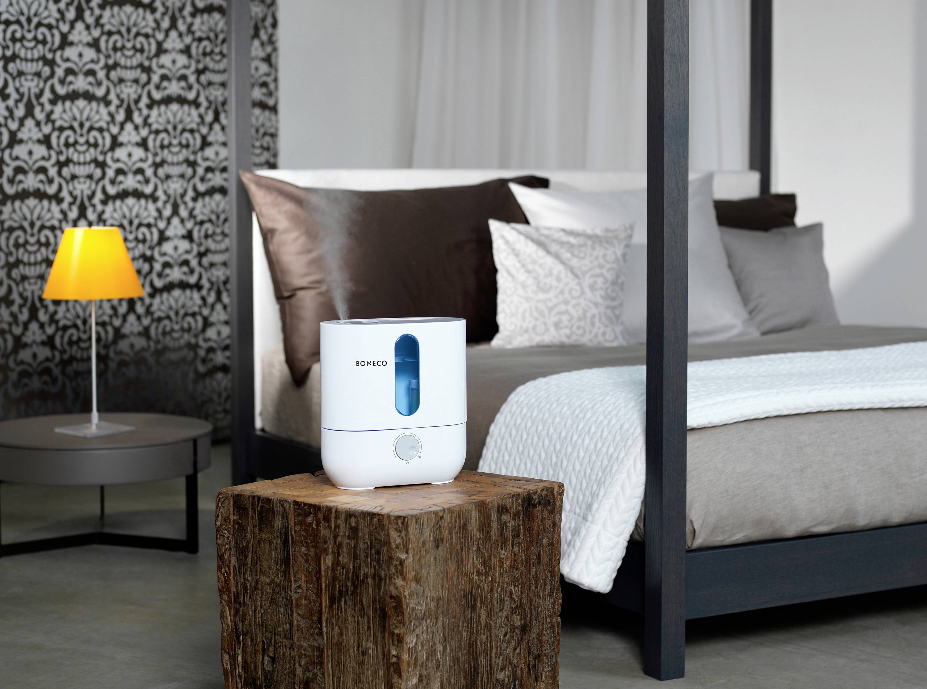 Un humidificateur est posé sur une table en bois devant un lit dans une chambre à coucher au design moderne. Une lampe brille en arrière-plan.