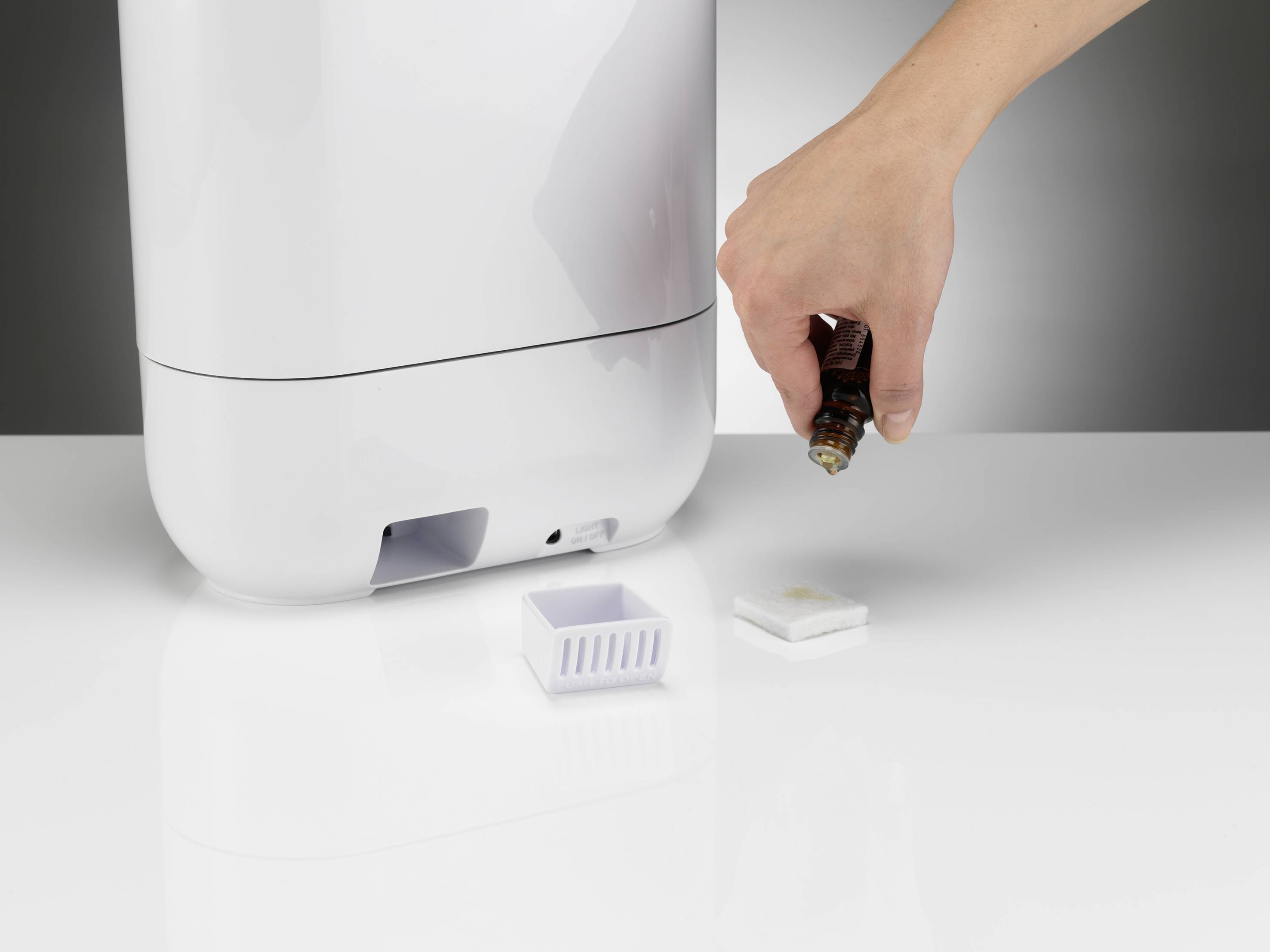 Une main fait tomber de l'huile essentielle sur un tampon près d'un humidificateur. Un petit compartiment est ouvert pour recevoir le tampon.