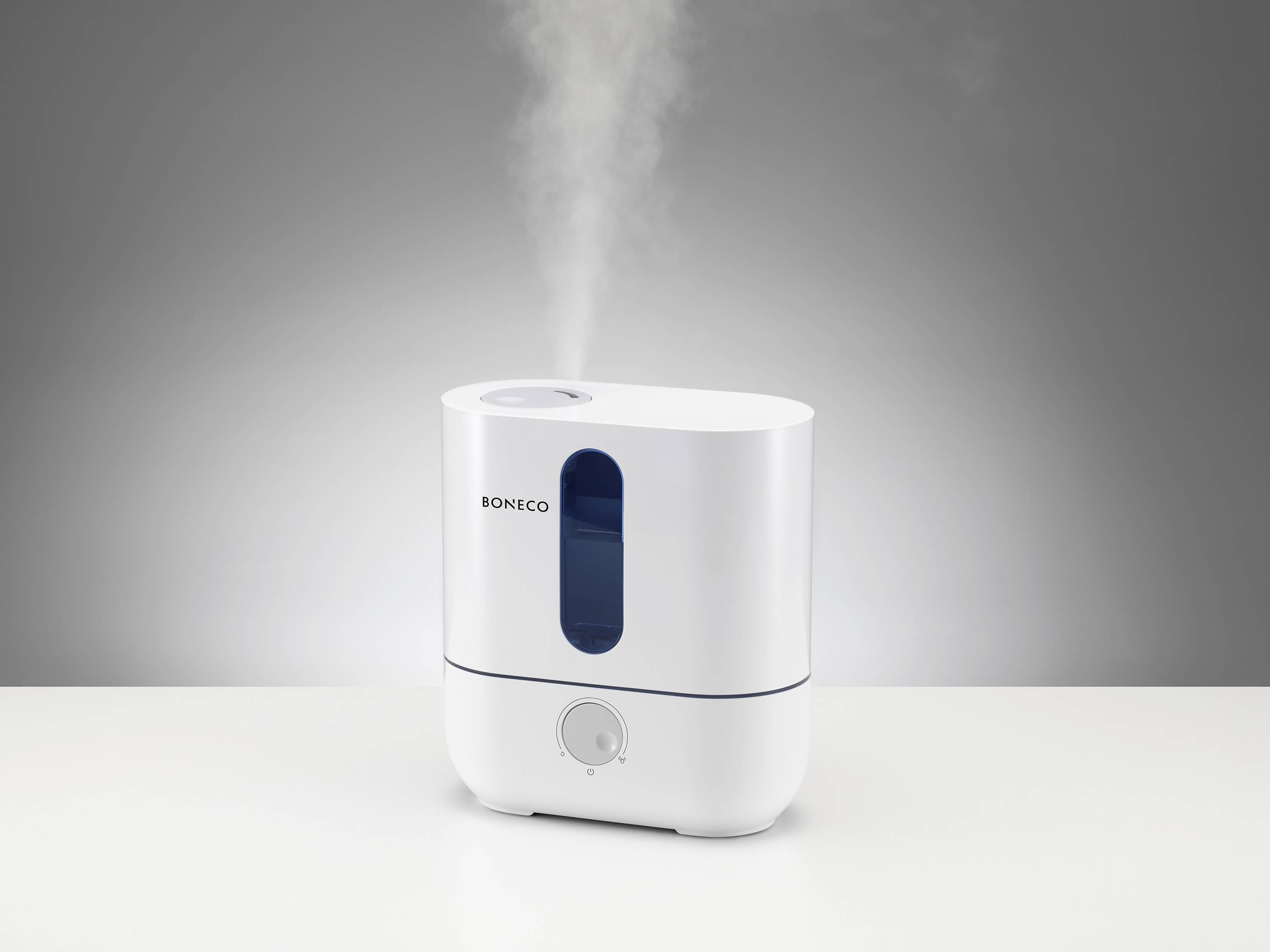 Un humidificateur blanc et rectangulaire, doté d'un indicateur de niveau d'eau bleu, diffuse de la vapeur vers le haut, posé sur une table.