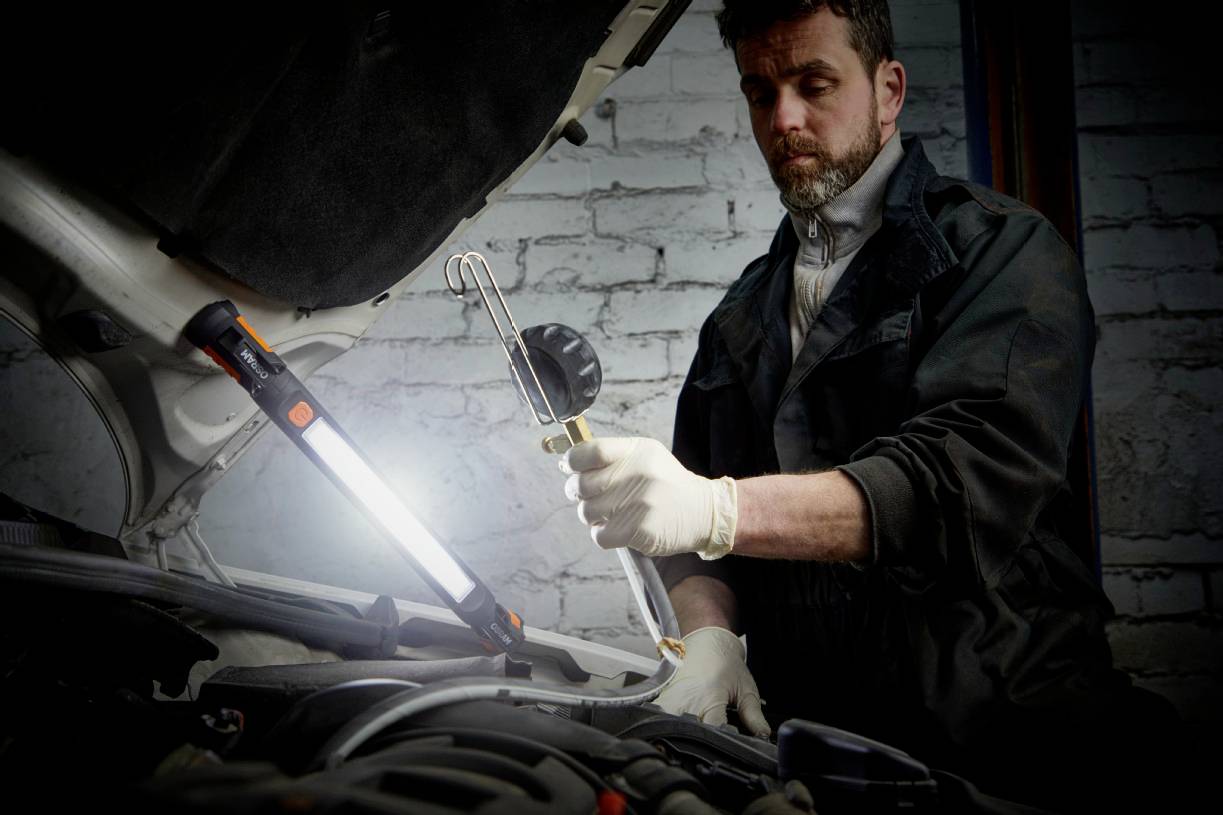 Un mécanicien vérifie le moteur d'une voiture dans un atelier. Il porte des gants et tient une lampe de travail dirigée vers le moteur.