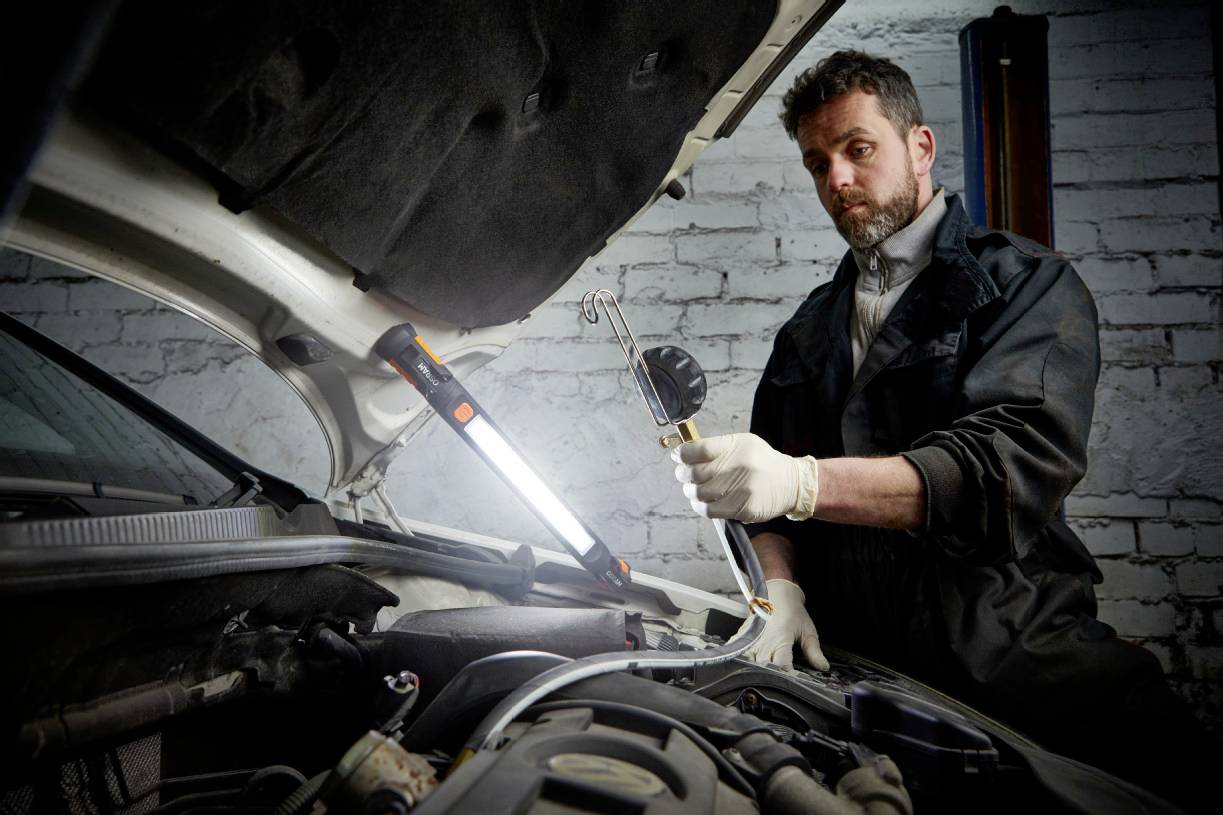 Un mécanicien répare un moteur, debout près d'une voiture ouverte avec une lampe de travail. Environnement d'atelier, mur de briques en arrière-plan.