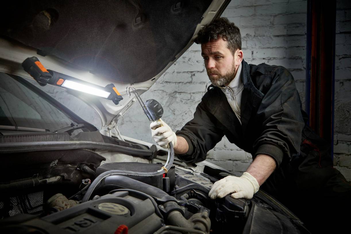 Un mécanicien en combinaison vérifie le moteur d'une voiture dans un atelier. Il tient un appareil de diagnostic, tandis qu'une lampe de travail éclaire la scène.