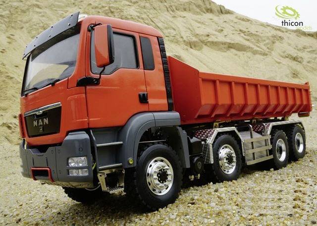 Un camion rouge est stationné sur un sol caillouteux devant une dune de sable. Un logo est visible en arrière-plan.