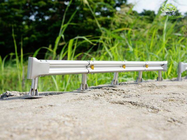 Une petite clôture de modèle en métal se dresse sur un sol pierreux, avec de l'herbe verte et de la végétation visible en arrière-plan.