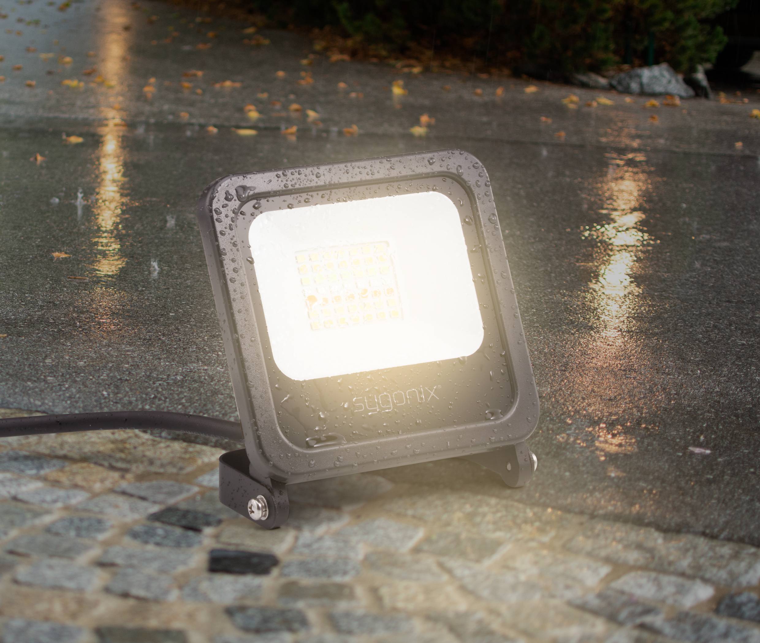 Un projecteur LED étanche s'illumine sur des pavés humides. La pluie se reflète sur le sol, l'éclairage souligne l'humidité.