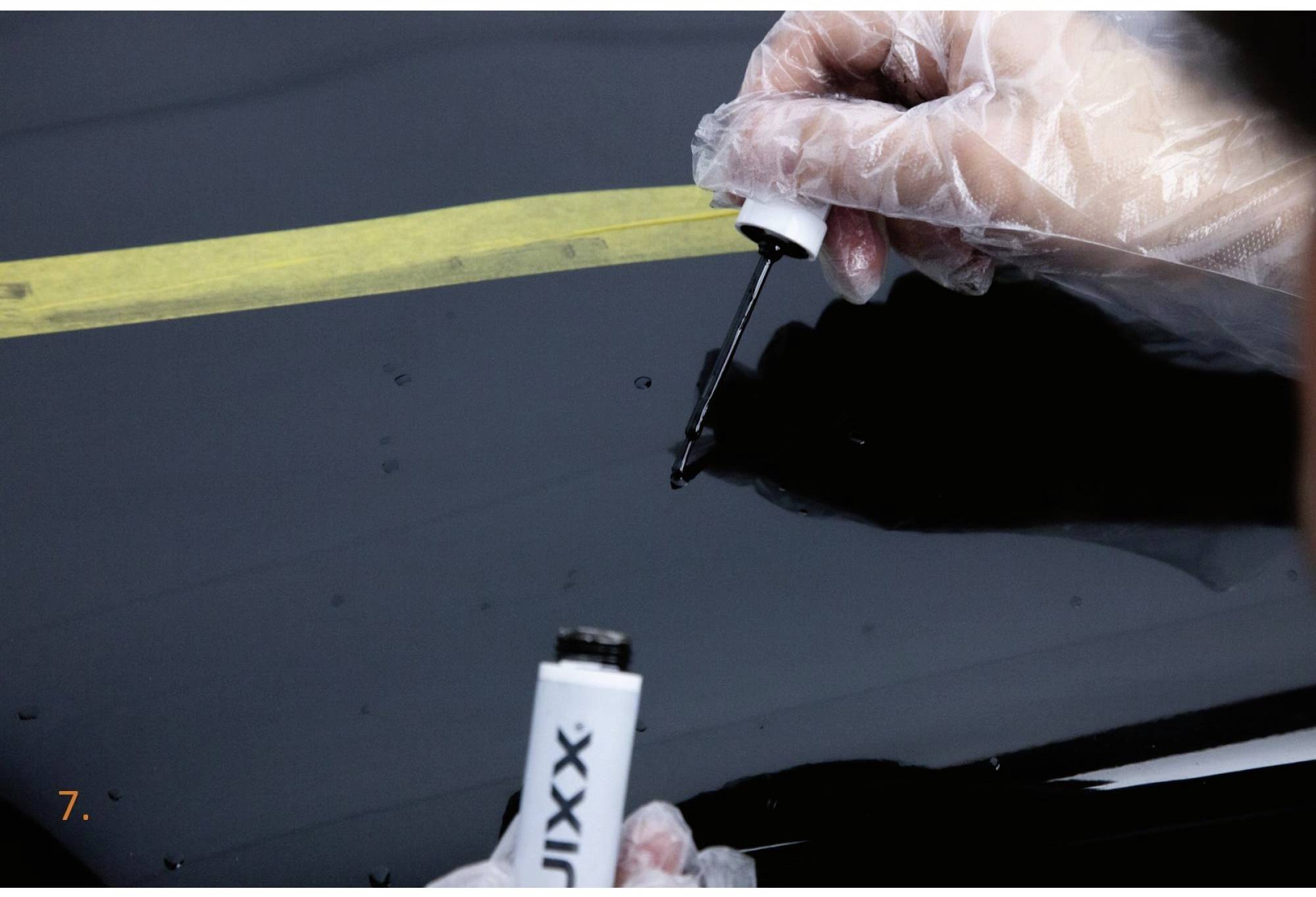 Une personne applique un liquide sur une rayure de surface sombre, en portant des gants et en utilisant un ruban adhésif jaune comme guide.