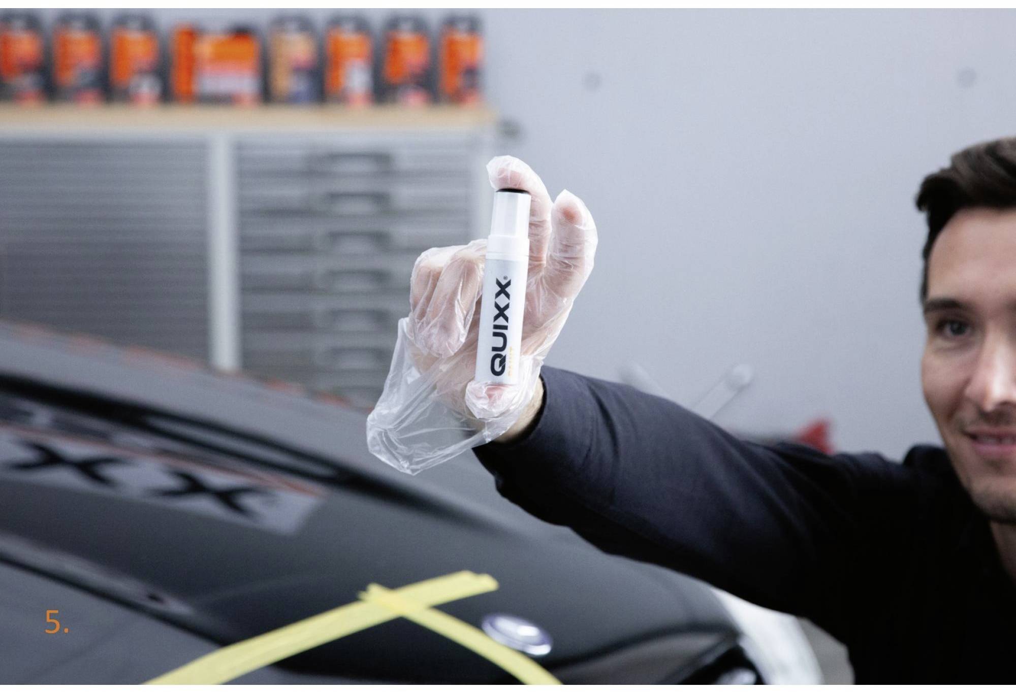 Un homme tient un stylo 'QUIXX' devant une voiture. Des produits d'entretien automobile sont visibles en arrière-plan.