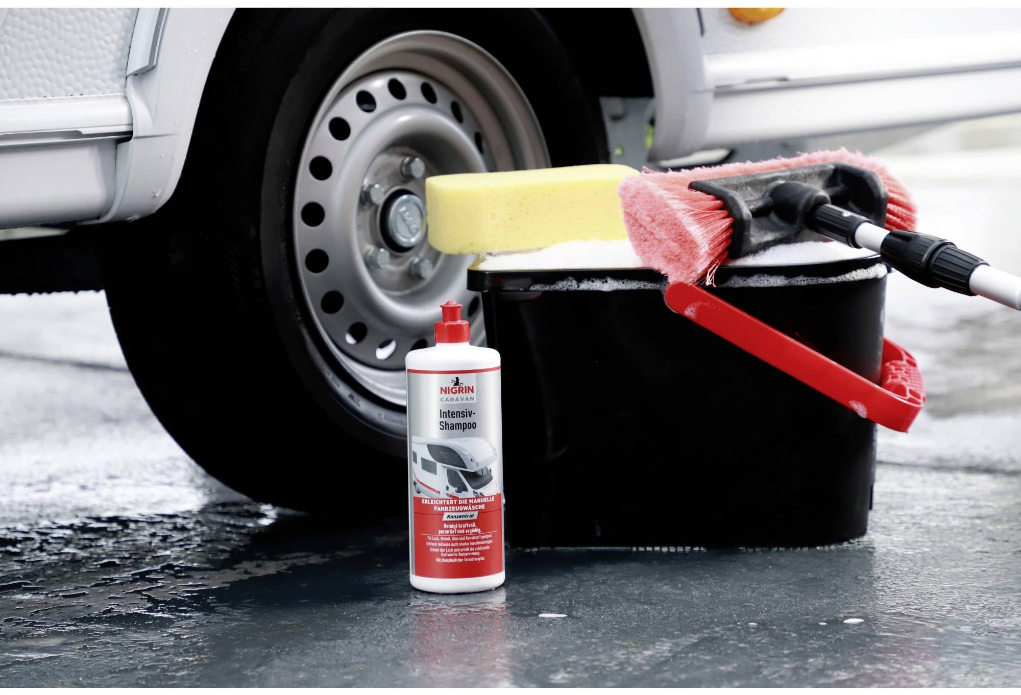 Une voiture est stationnée sur un asphalte humide. À côté, un seau avec une éponge et une brosse, ainsi qu'une bouteille de shampoing pour voiture. La scène représente des accessoires d'entretien automobile.