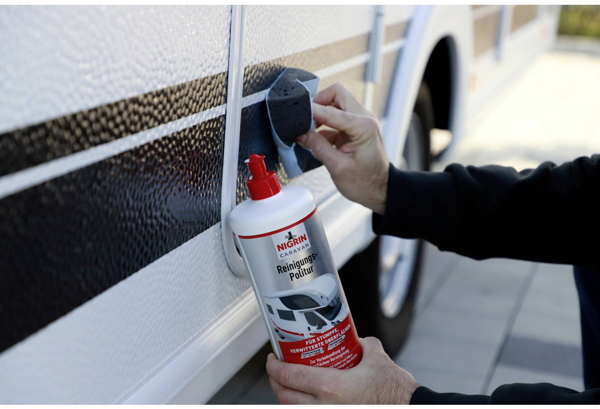 Une personne nettoie une caravane blanche avec un produit de nettoyage et de polissage. Une bouteille rouge et une éponge sont utilisées.