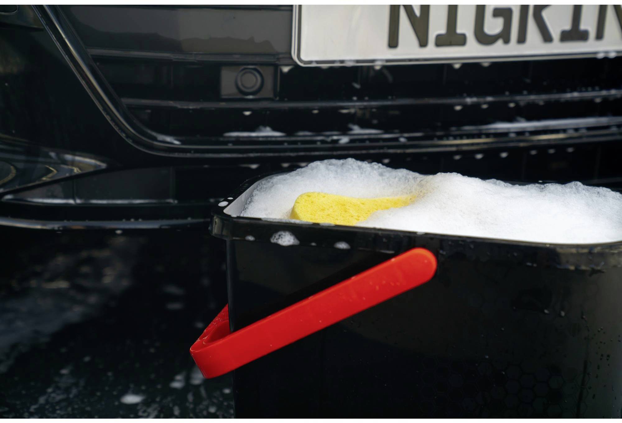 Un seau noir rempli d'eau savonneuse et une éponge jaune sont placés devant une voiture noire dont une partie de la plaque d'immatriculation est visible.