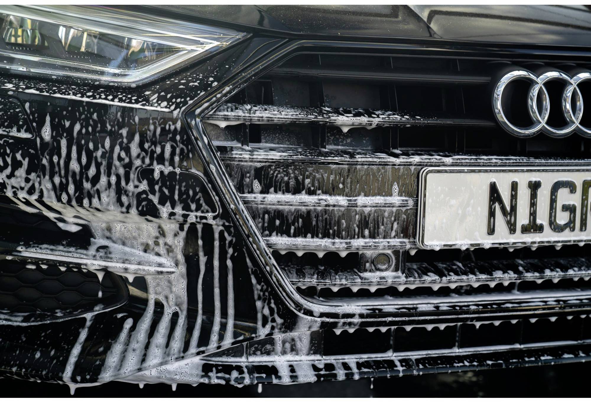 En gros plan, l'image montre une Audi noire lors d'un lavage automobile, avec un focus sur la calandre et la plaque d'immatriculation.
