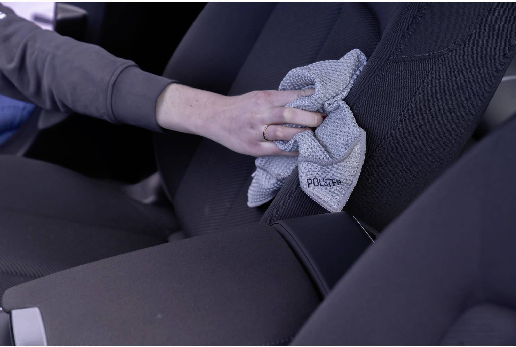 Une personne nettoie le siège d'une voiture avec un chiffon gris. Elle porte un haut à manches longues. L'accent est mis sur l'action de nettoyage.
