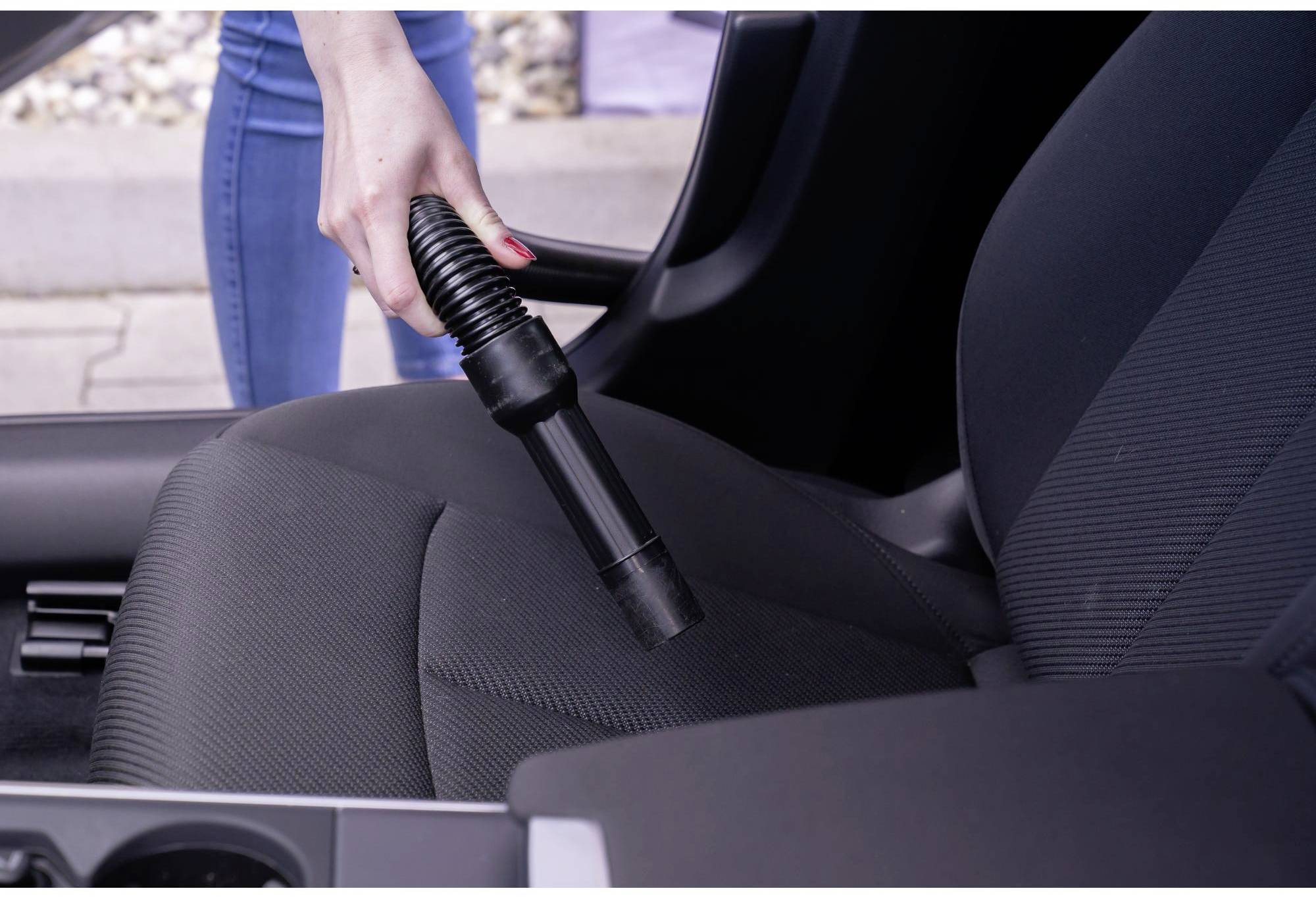 Une personne aspire le siège d'une voiture avec un aspirateur. Le focus est mis sur la main qui tient le tuyau de l'aspirateur.