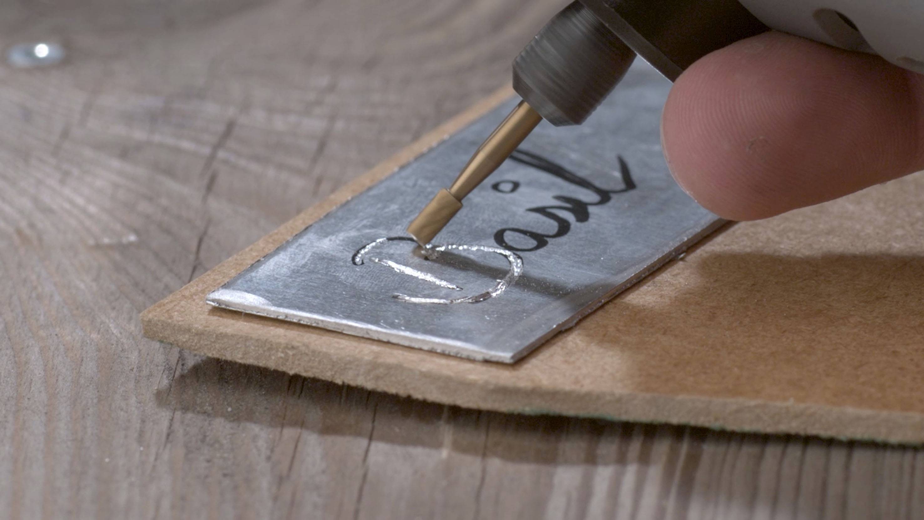 Une main tient un stylet de gravure et grave le mot 'Basil' sur une plaque de métal posée sur une table en bois.
