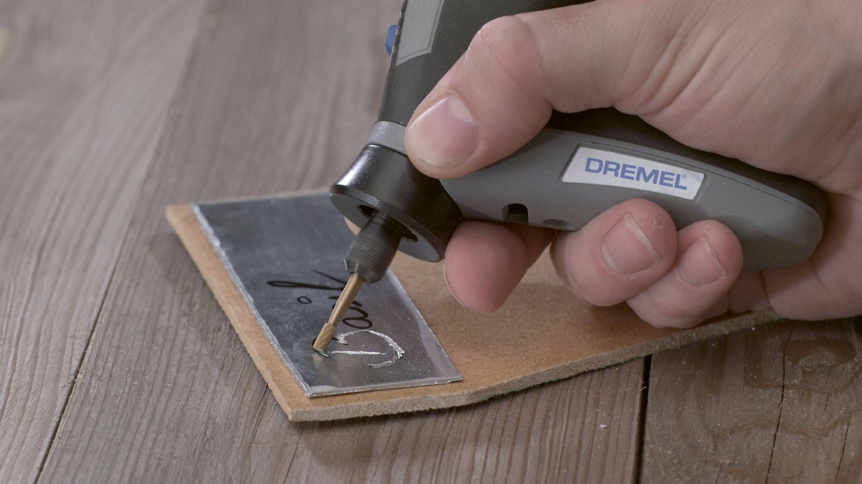 Une main tient un outil de gravure électrique avec lequel le nom 'David' est gravé sur une plaque métallique posée sur une table en bois.