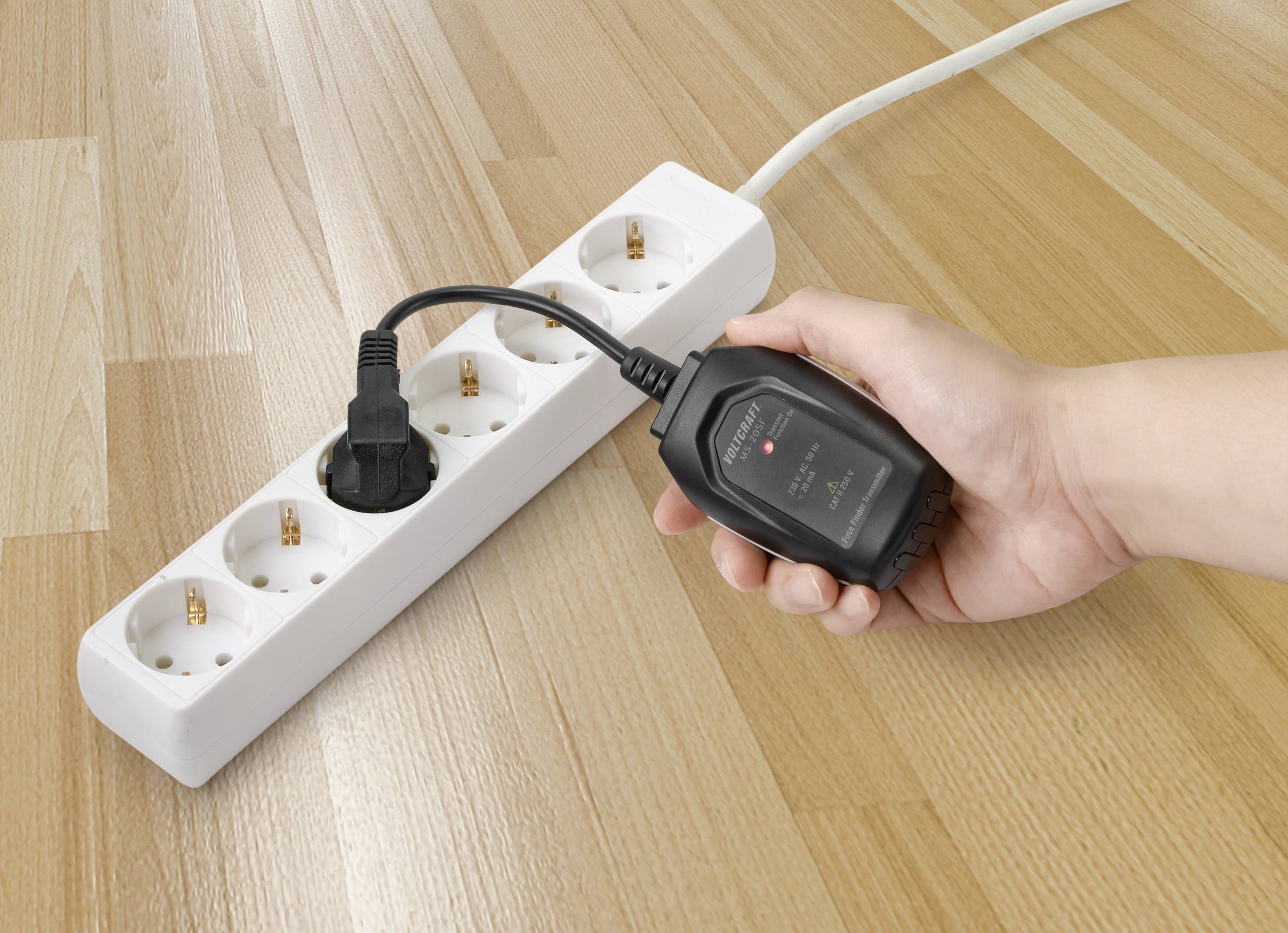 Une main tient un appareil de mesure électrique près d'un câble électrique branché dans une multiprise blanche pour vérifier le flux de courant.