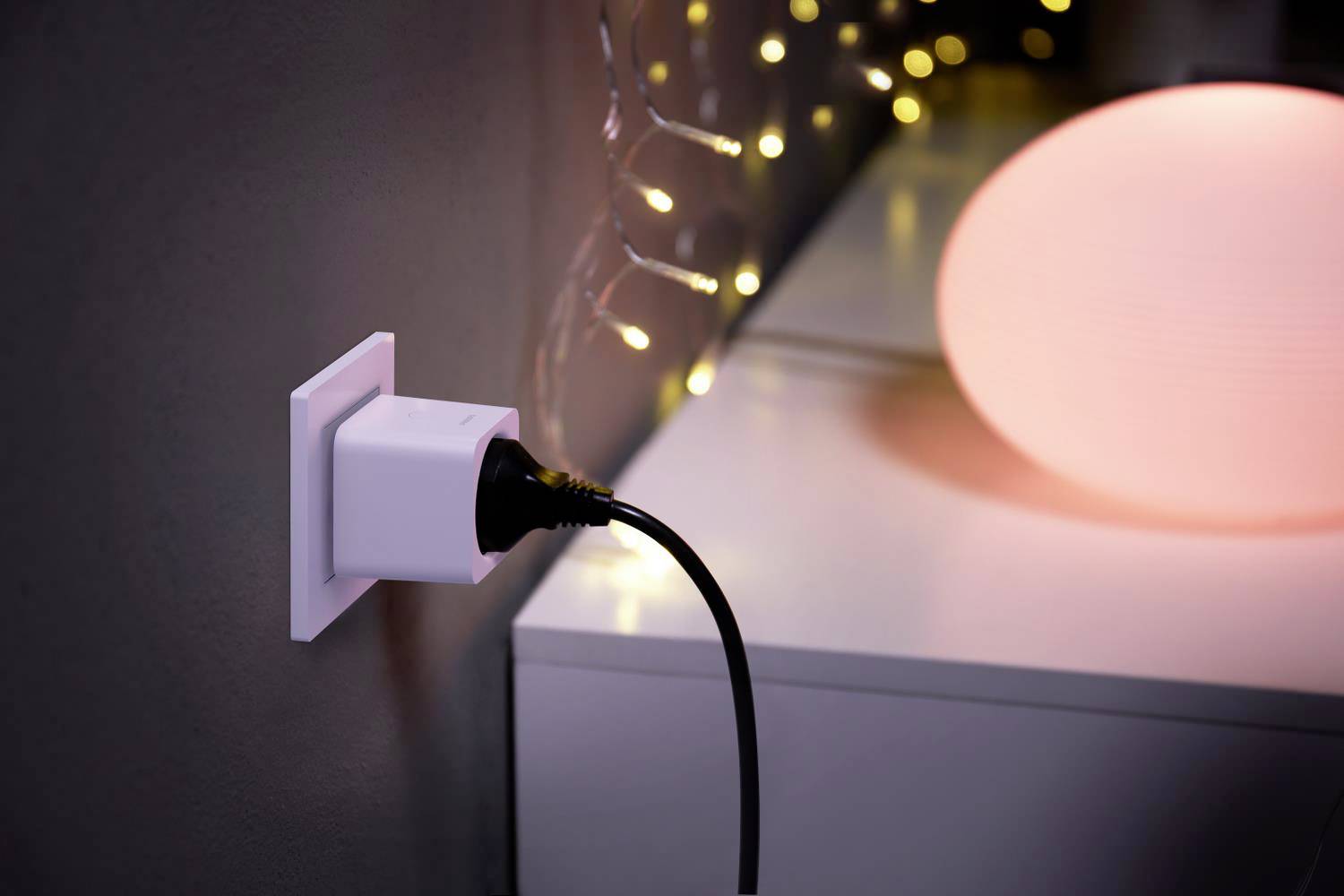 Une prise électrique avec un câble noir branché. À côté, une table avec une lampe allumée et des guirlandes lumineuses en arrière-plan.