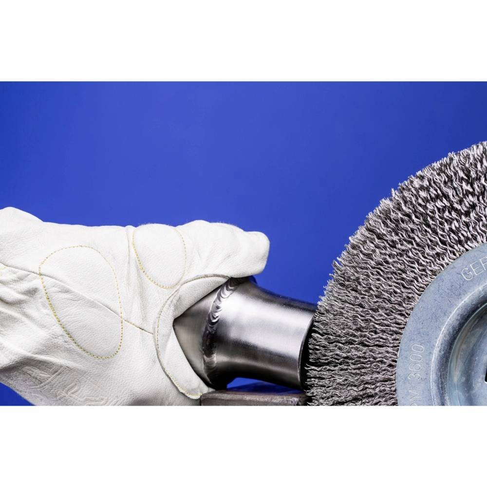 Une main protégée par un gant blanc tient une pièce métallique contre une brosse métallique rotative sur un fond bleu.
