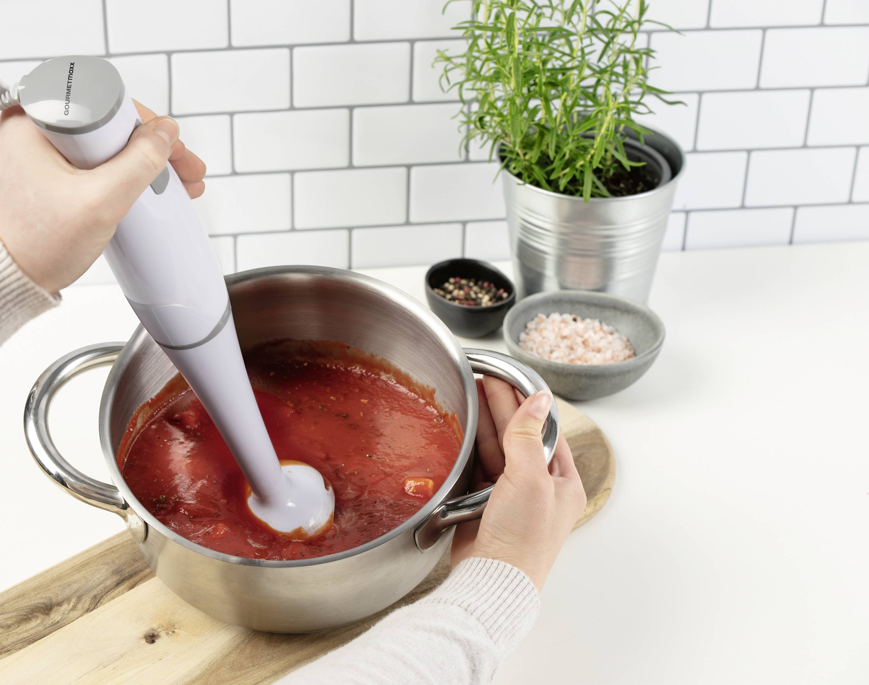 Une personne mixe une sauce rouge avec un mixeur plongeant dans une casserole en acier inoxydable. Des épices et une plante sont visibles en arrière-plan.