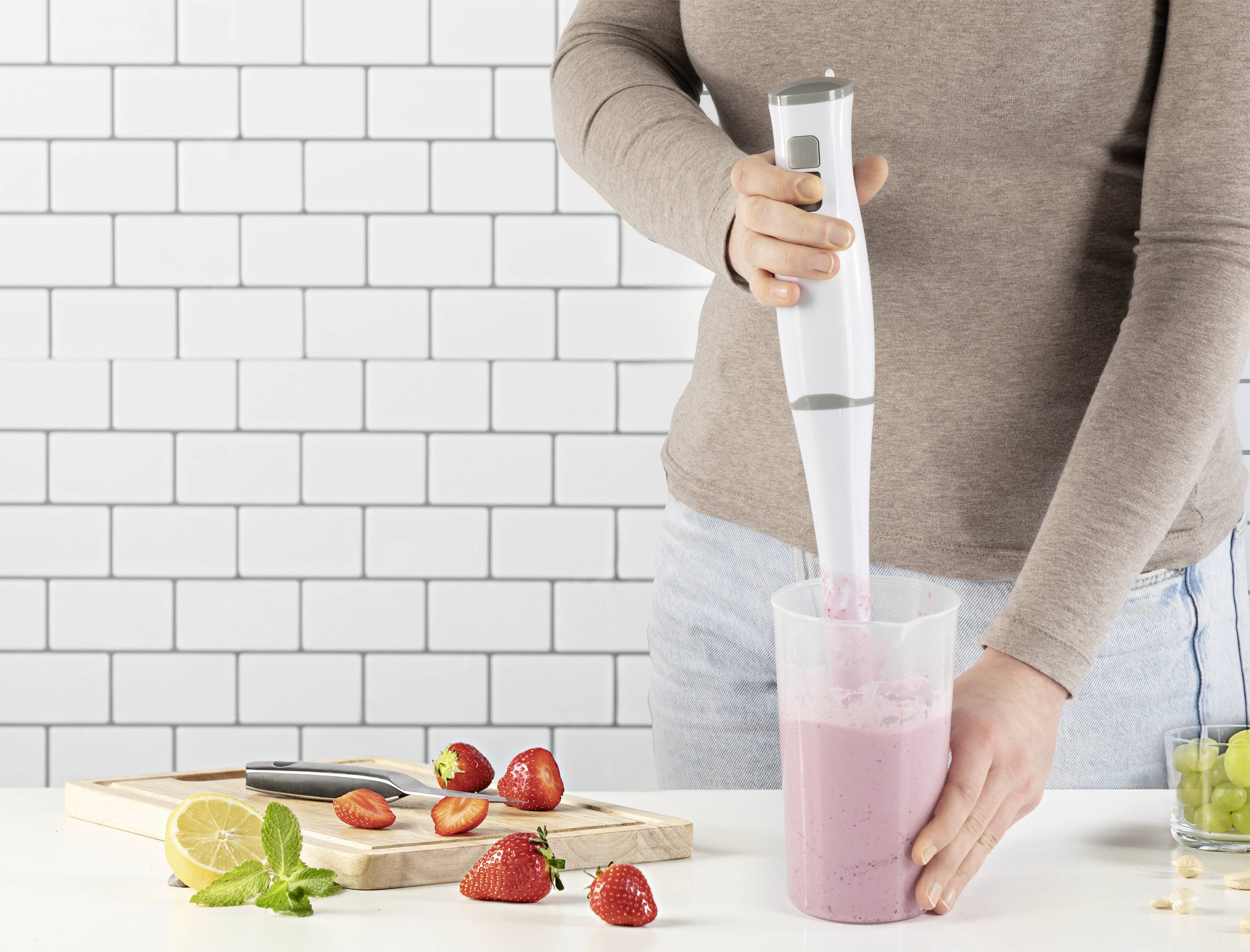 Une personne mixe un smoothie aux fraises avec un mixeur dans un récipient doseur. Sur la table sont posés des fraises et des tranches de citron.