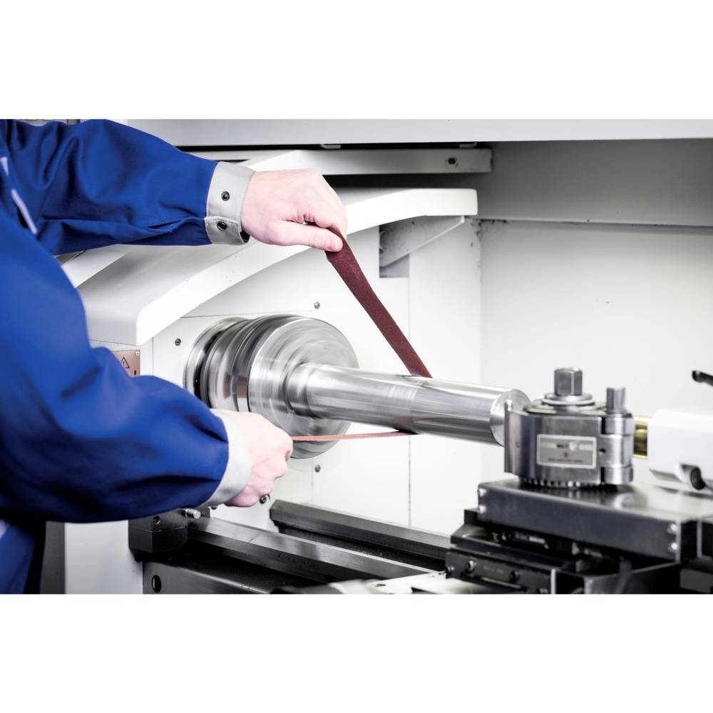 Un ouvrier métallurgiste travaille une tige métallique cylindrique sur une machine équipée d'un outil de meulage dans un atelier.