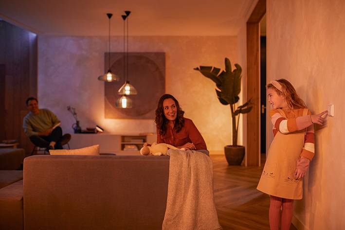 Un enfant allume la lumière dans un salon doucement éclairé, tandis qu'une femme est assise sur le canapé et sourit.