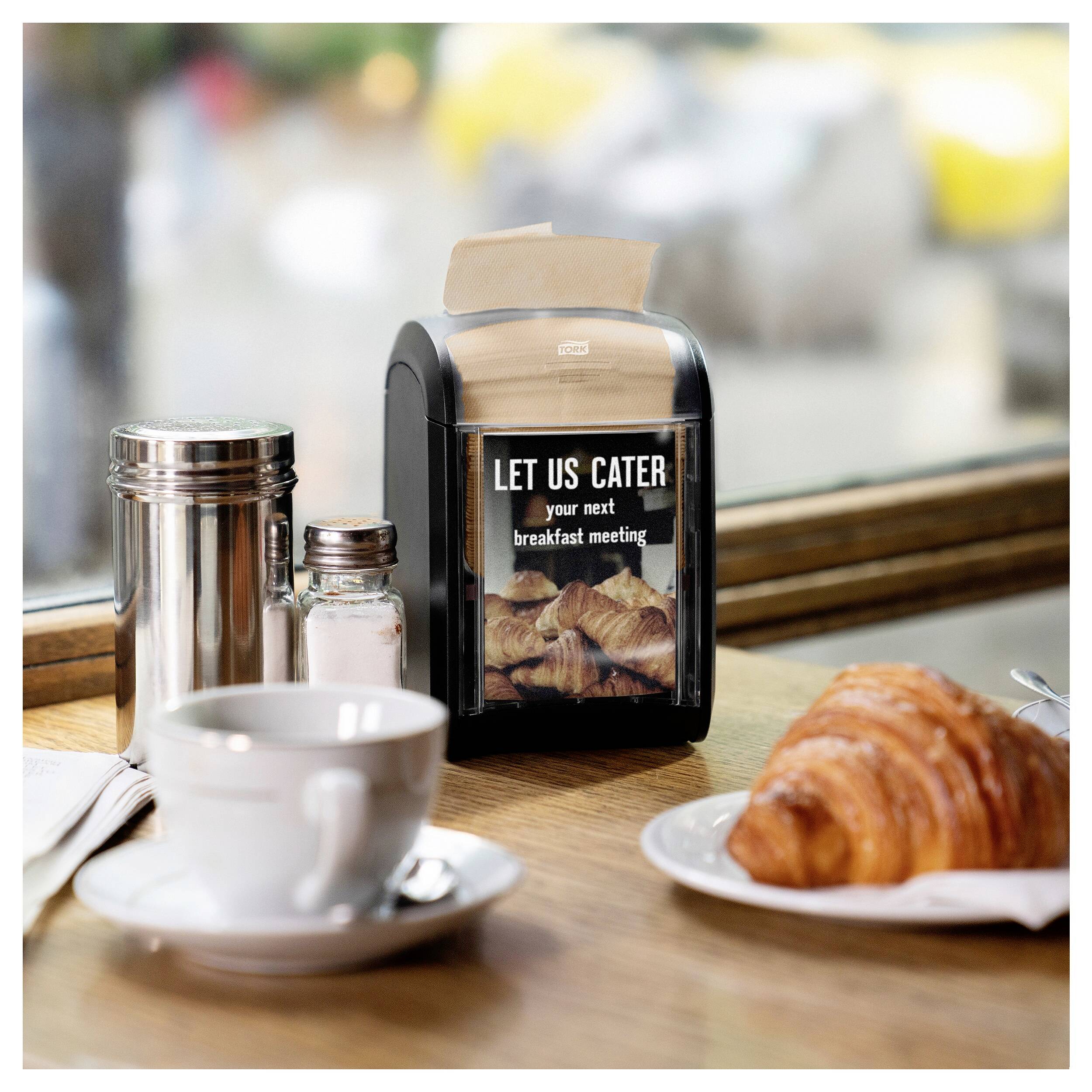 Sur une table, il y a une tasse, un croissant et un distributeur de papier portant l'inscription « Laissez-nous organiser votre prochain petit-déjeuner de réunion ».