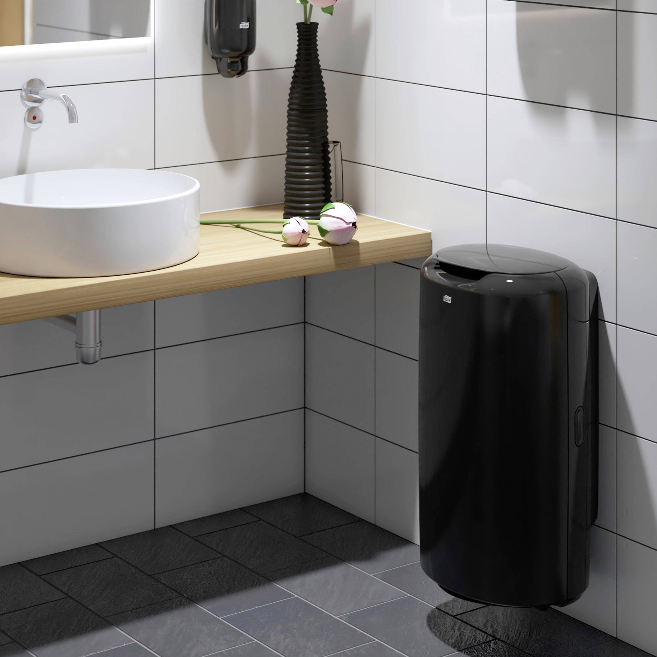 Une salle de bain moderne avec un lavabo sur un plateau en bois, un vase de fleurs posé dessus et une poubelle noire à côté, le tout dans des tons clairs.