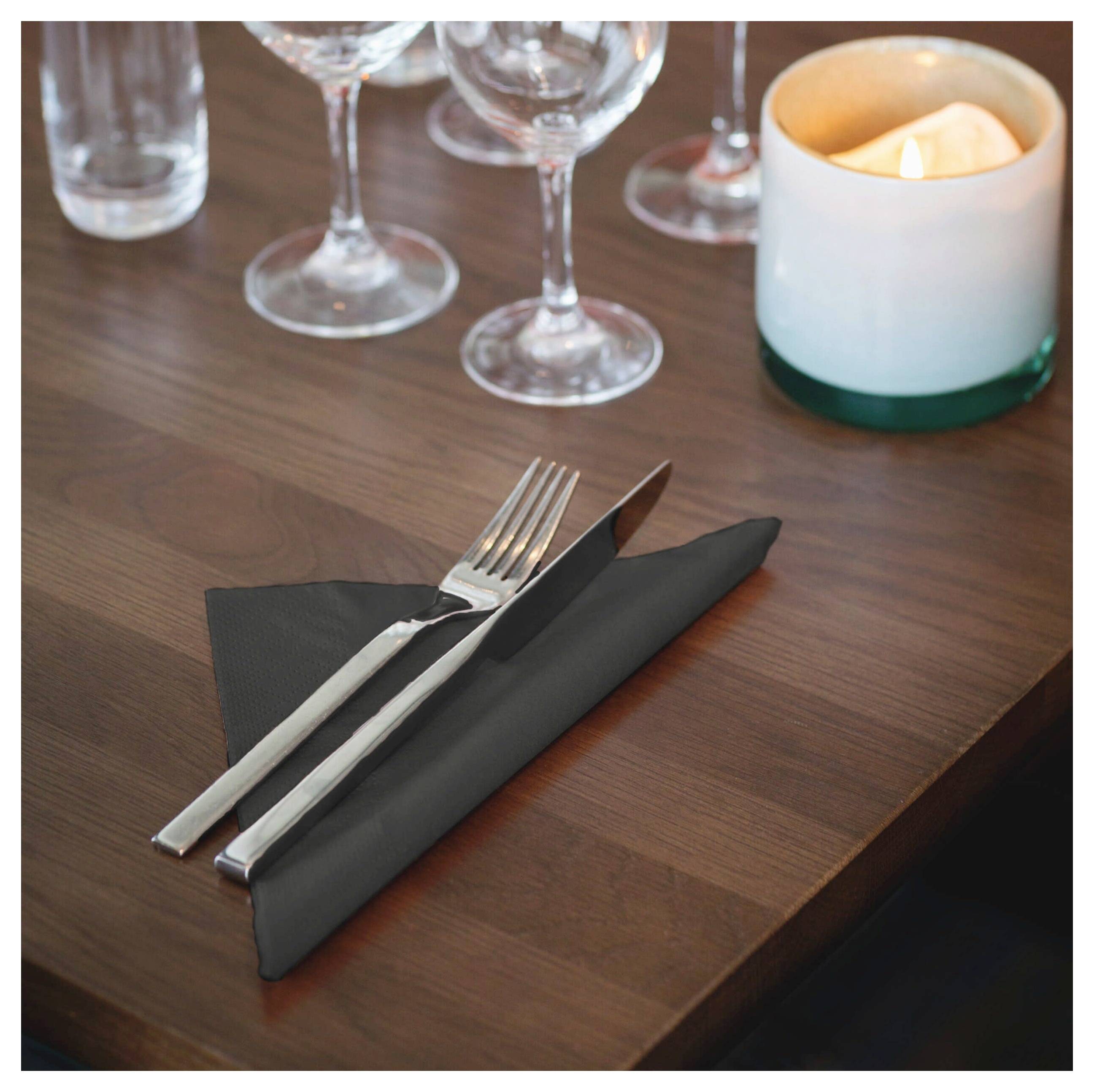 Gedeck mit gefalteter schwarzer Serviette, Gabel und Messer, Weingläsern und einer brennenden Kerze auf einem Holztisch.