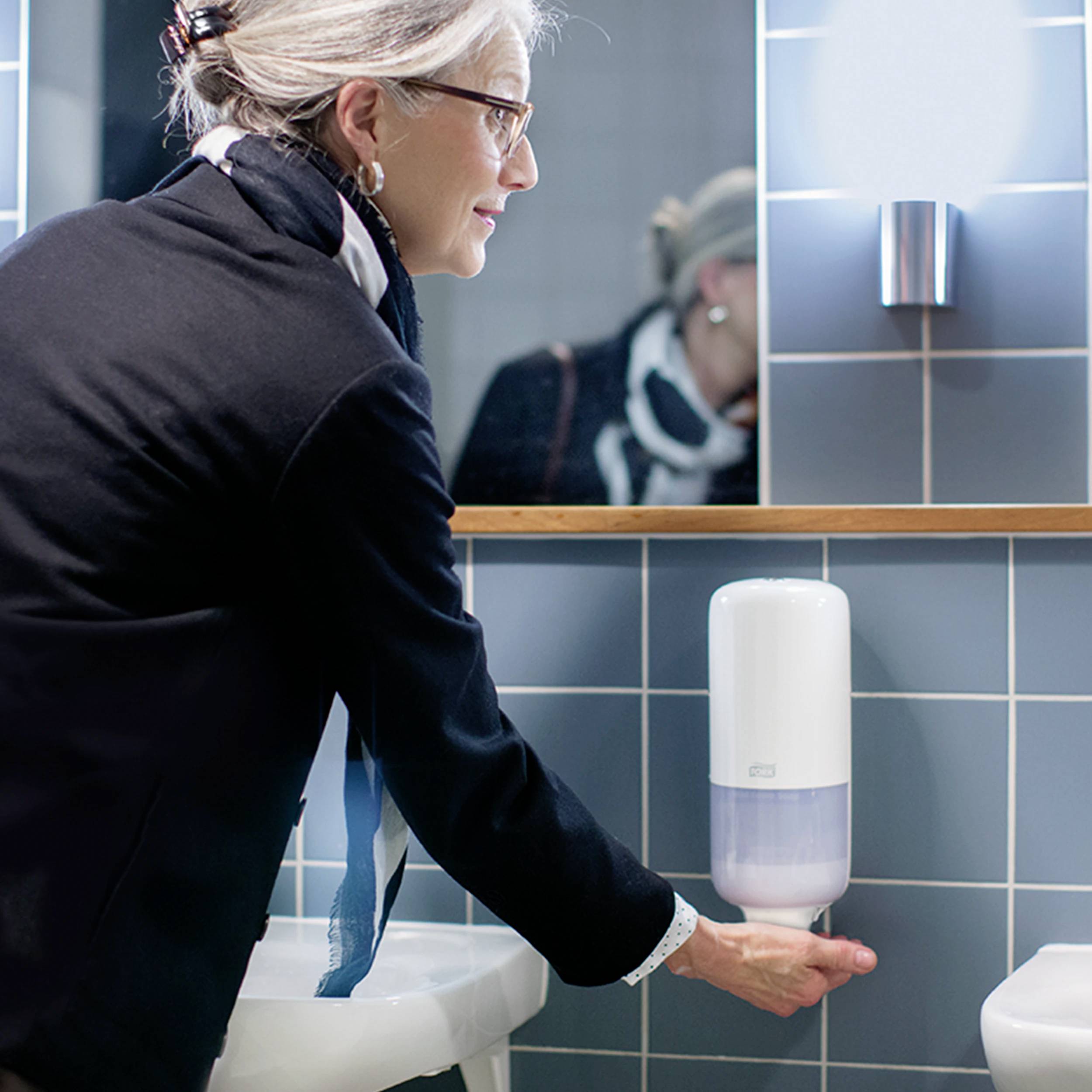 Une personne appuie sur un distributeur de savon mural dans une salle de bain. Des carreaux bleus sont visibles en arrière-plan.
